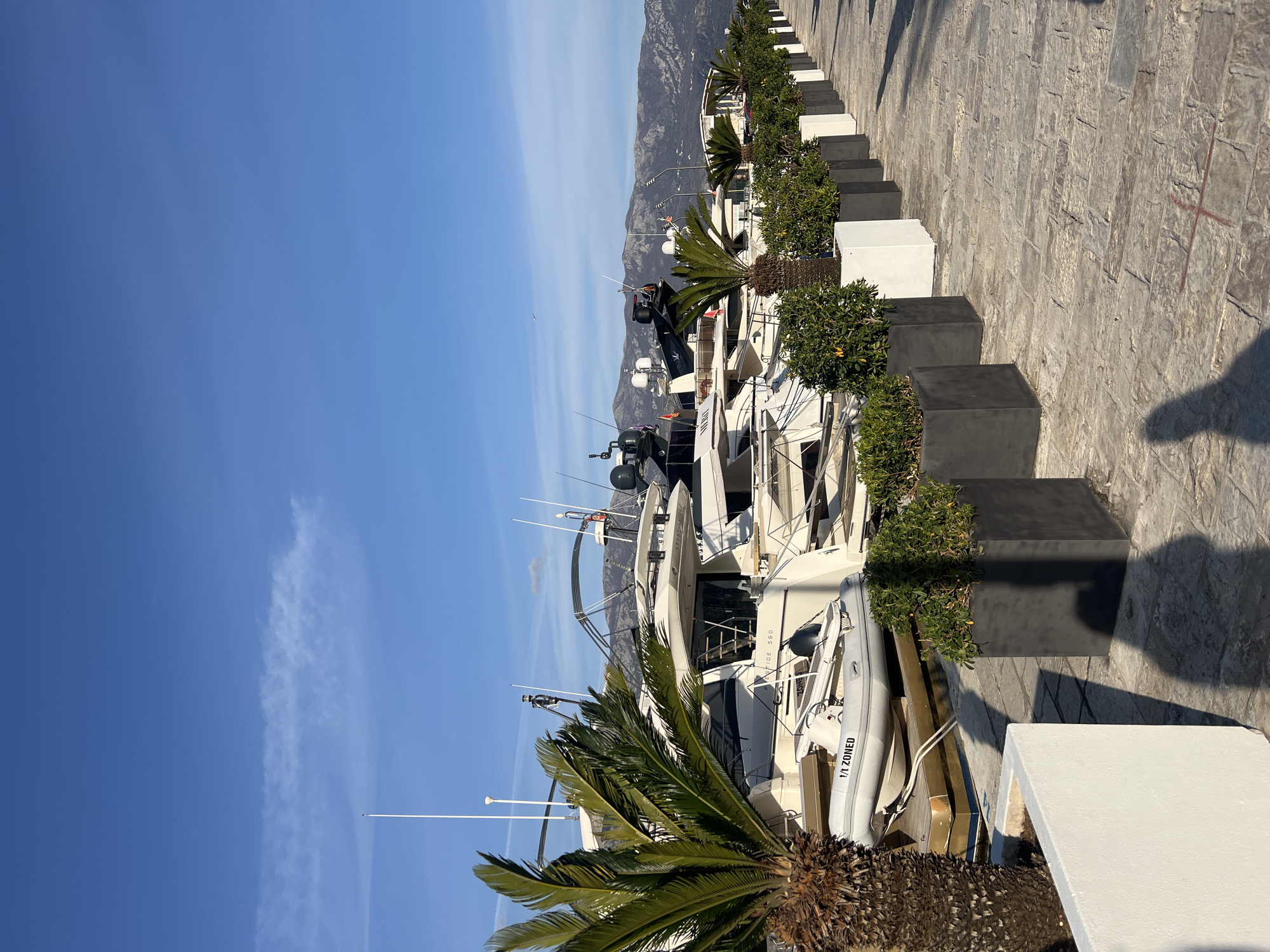 The image depicts a marina scene in Budva, Montenegro, under a clear, bright blue sky with sparse, wispy white clouds, including a long, horizontal cloud formation in the upper sky. In the background, rugged mountains are visible under the horizon line, partially obscured by numerous docked vessels.
The midground is dominated by a series of luxury yachts and motor cruisers, predominantly white and dark-colored, featuring multiple decks, antennas, radar domes, and railings. One visible vessel is labeled "PRESTIGE 590". Several smaller, grey inflatable dinghies or tenders are stowed on the larger yachts or directly on the dock, one of which bears the text "1/1 ZONED". The boats are moored parallel to a paved promenade.
The promenade, located in the foreground and extending into the midground, is constructed from irregular grey stone blocks. A line of planters runs parallel to the boats. These planters alternate between white and dark grey rectangular shapes, containing green bushy plants and several Sago palm trees (Cycas revoluta) with visible brown trunks. Closer to the viewer, a larger, white concrete low planter is partially visible.
No people are explicitly visible, but a distinct elongated shadow of an individual, likely the photographer, is cast across the paved promenade and a white planter in the foreground, indicating the presence of a person. A red 'X' mark is painted directly on the grey stone pavement in the lower right foreground. Strong sunlight illuminates the scene, casting sharp shadows from the plants and planters.