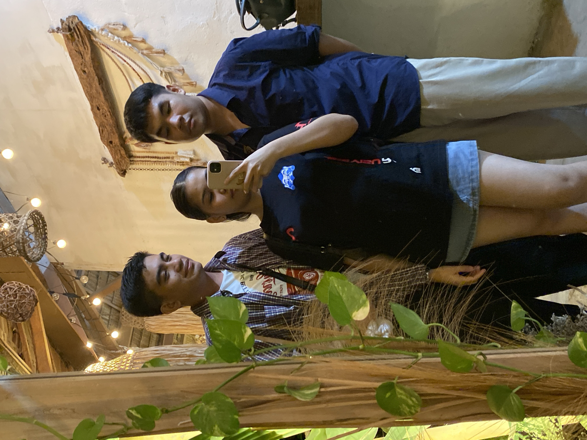 Three young adults are posing for a mirror selfie in what appears to be an outdoor dining or lounging area. The person in the center, a young woman, is holding a smartphone to take the picture. She is wearing a black t-shirt and a denim skirt. To her left is a young man wearing a plaid shirt over a white t-shirt. To her right is another young man wearing a dark blue collared shirt and light-colored pants. The background features a rustic ceiling with exposed wooden beams and hanging decorative lights. There's also a mirror, a wooden beam adorned with natural materials, and greenery from plants. The overall atmosphere suggests a casual and tropical setting, likely in Samal, Philippines, given the mention of the location.