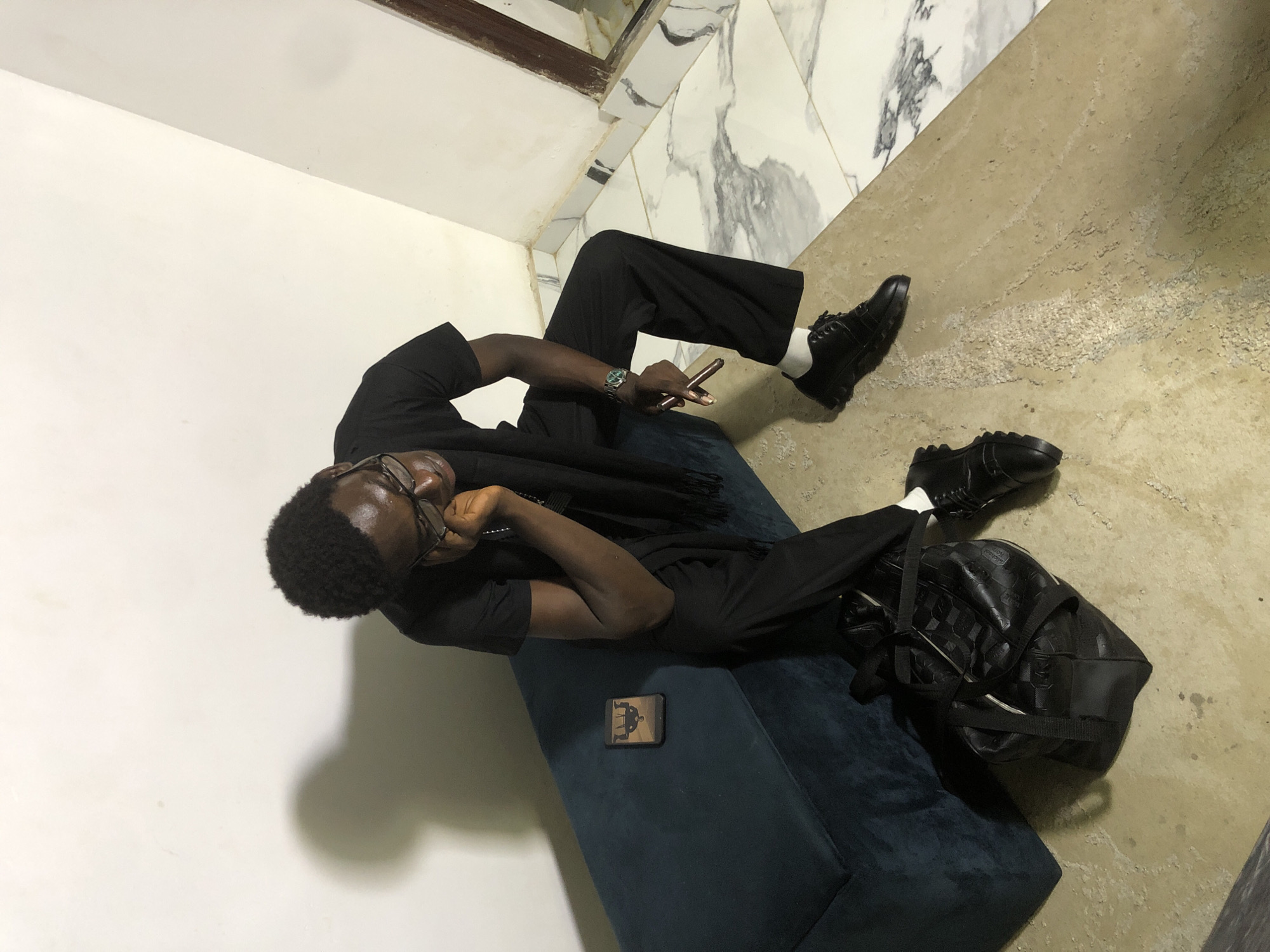 A young Black man wearing glasses, a black t-shirt, black pants, and black shoes is sitting on a dark blue bench in what appears to be a well-lit indoor space with white walls and marble flooring. He is leaning his head on his hand, with his eyes closed or looking down. Next to him on the bench is a smartphone with a cartoon image displayed on its screen. On the floor in front of the bench are a black duffel bag and another pair of black shoes. The man is wearing a watch on his left wrist and is holding a thin, cylindrical object in his right hand. The overall scene suggests a moment of quiet contemplation or rest in a modern setting. The location is Ado, Nigeria.