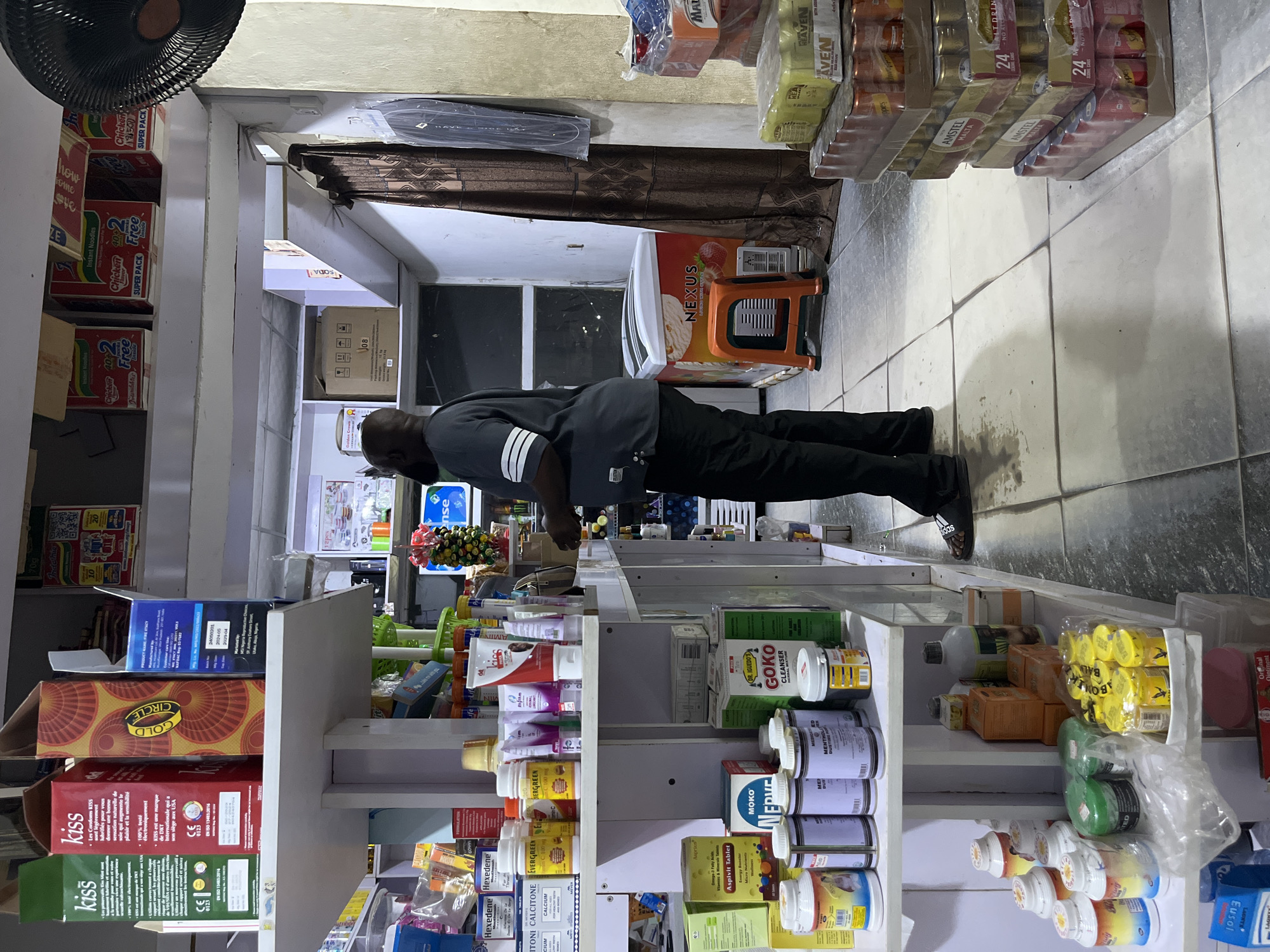 A person is doing push-ups in a small store. The store sells various items including condoms, medication, and instant noodles. The location is in Abuja, Nigeria.