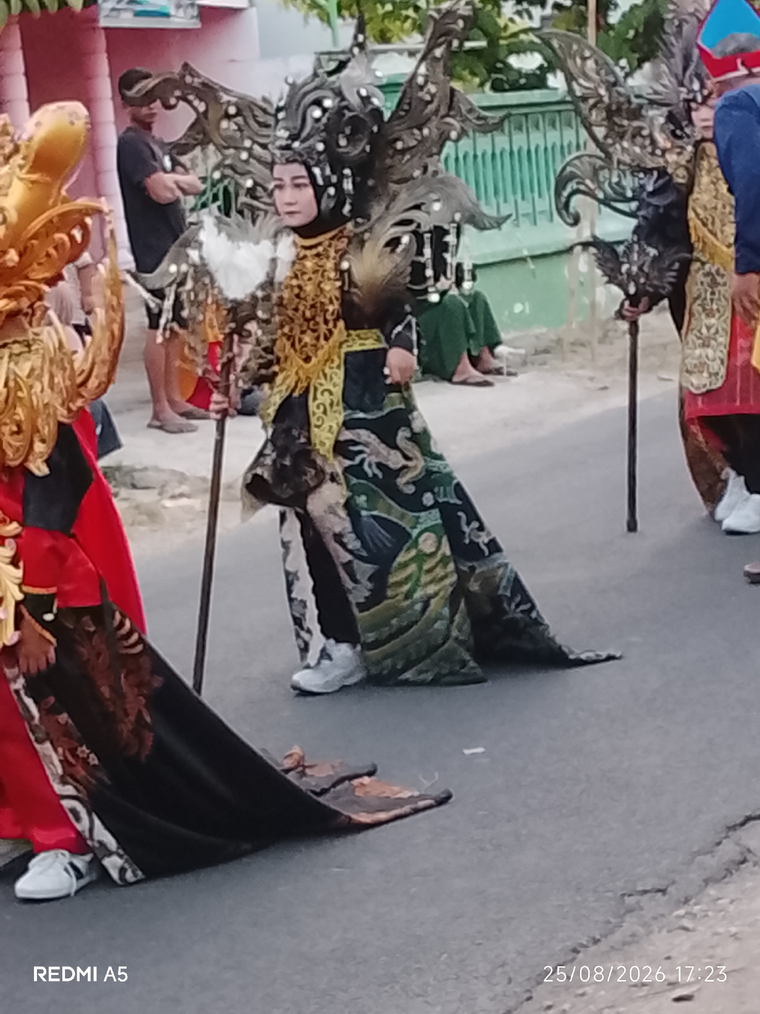 In Semanding, Indonesia, a cultural parade is underway on a paved road. The foreground features two individuals in elaborate costumes. The person on the right wears a headdress with black, spiky protrusions and a detailed gold and black patterned garment with a long train. They hold a dark pole. To their left, another person is adorned in a gold dragon-like headpiece and a red garment, also with a trailing fabric. In the background, several other people are visible. One individual in a dark t-shirt and shorts stands with arms crossed. Others are seated or standing further back, some wearing green clothing. The environment consists of a road, a light-colored building on the left, and a green fence in the background with trees beyond. The overall scene depicts a festive, outdoor cultural event.