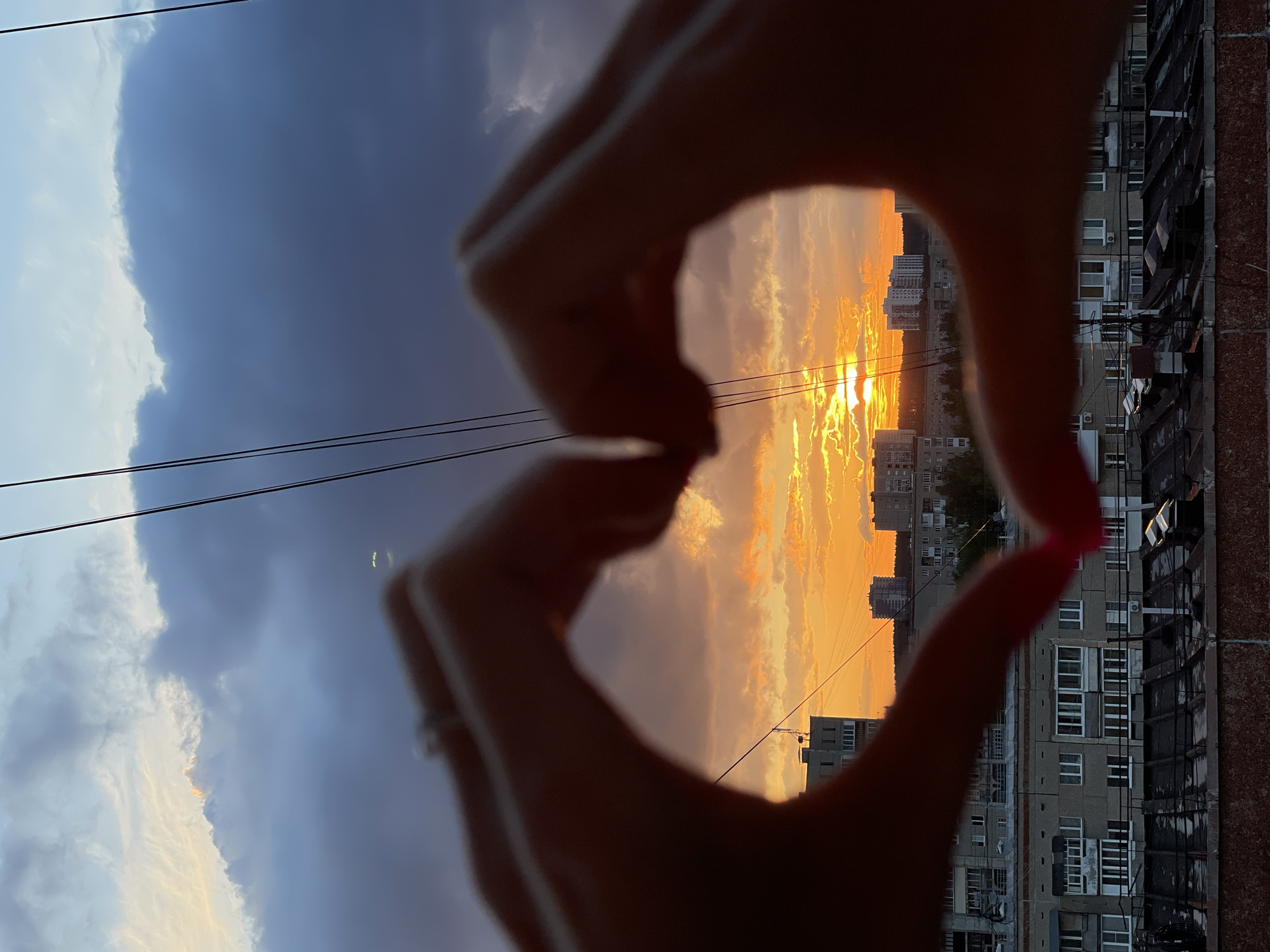 A view of a sunset through a heart-shaped frame made by hands in Izhevsk, Russia. The sky is a mix of dark and light clouds, with the sun setting behind buildings. Apartment buildings are visible in the background. Overhead power lines are in view. One fingernail has red polish.