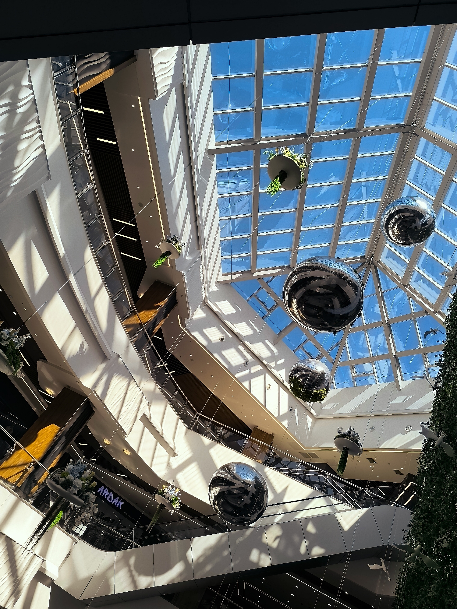 The image depicts the interior of a modern shopping mall, likely located in Qarağandı city administration, Kazakhstan, based on a visible sign. The viewpoint is from a high angle, looking upwards towards a large, glass-paned atrium roof. Sunlight streams through the roof, casting geometric shadows across the multi-level interior.
The mall features multiple floors with escalators and walkways. The architecture is sleek and contemporary, with white, curved walls and dark accents. Suspended from the ceiling are several large, reflective spherical ornaments, some containing greenery, and decorative floral arrangements. A large, conical structure resembling a Christmas tree, adorned with white bird-like ornaments, is visible on the right side of the frame. The overall impression is one of a bright, spacious, and festive commercial environment. No people are visible in the image.