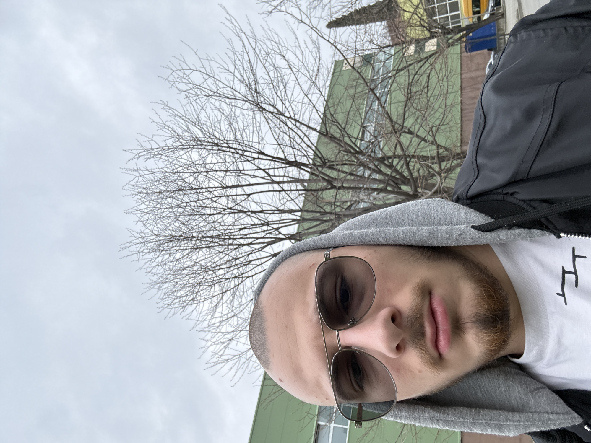 A close-up selfie depicts a male subject outdoors in Stara Zagora, Bulgaria, under an overcast grey sky. The subject, positioned centrally, has a closely shaved head and a light brown/ginger stubble beard and mustache. He wears aviator-style sunglasses with dark lenses and thin metal frames. His attire consists of a grey hooded sweatshirt with the hood up, a white t-shirt featuring a dark, abstract symbol resembling two connected 'T' shapes on the chest, and a black jacket.
Behind the subject, a bare deciduous tree with numerous fine branches occupies the mid-ground. Further back and to the left, a large, light green industrial-style building with multiple horizontal windowpanes is visible. To the right of the tree, portions of other urban elements are present: a blue portable toilet, a yellow vehicle (likely a van or bus), and a dark-colored parked car on what appears to be a paved area or street. Another building section with windows is partially visible to the far right. The overall impression is an urban setting during a cooler season.