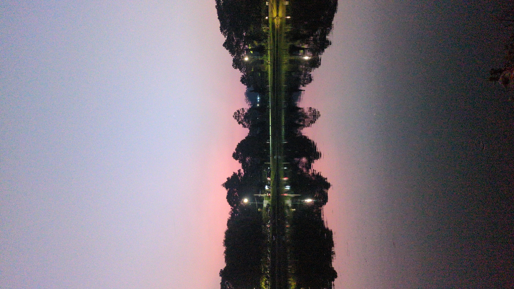 The image shows a reflection of a street or path in a body of water, likely a lake or pond. The street is lined with trees and streetlights. The sky above is a gradient of colors, ranging from light purple to pink, suggesting a sunset or sunrise. The water's surface is slightly disturbed, creating a rippled reflection. The location is possibly in Da Lat, Vietnam. There are no people immediately visible. The main subjects are the street, trees, water, and sky. The action is reflection, and the environment is a tranquil, possibly urban, park setting at dusk or dawn.