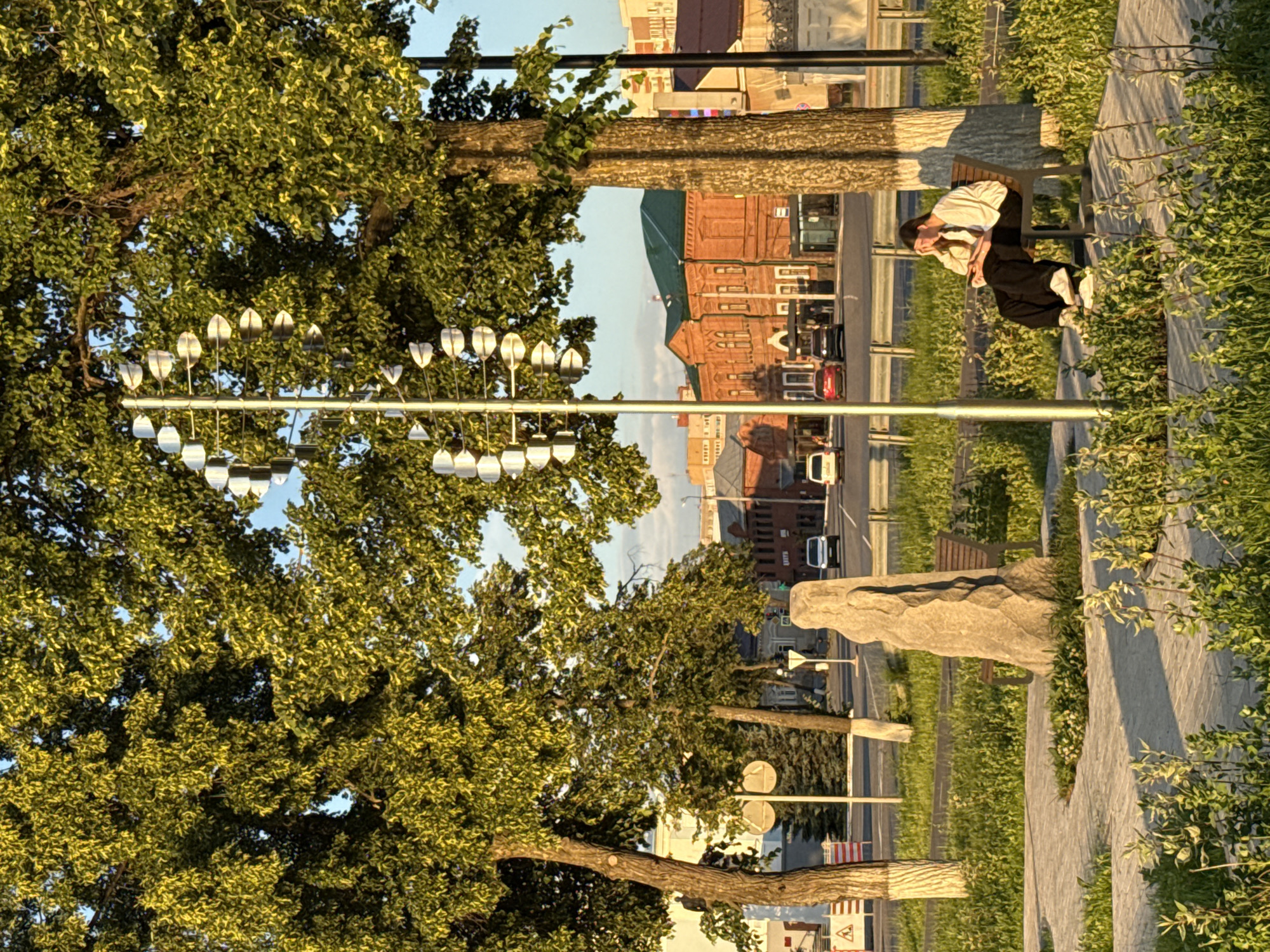 A young woman with dark hair, wearing a cream-colored sweatshirt and black pants, sits on a wooden bench in a park. She is looking down at her phone, with her left hand resting on her forehead. The park is illuminated by the warm, golden light of the setting sun. Lush green grass and various plants surround the bench and a paved walkway. In the background, a tall, silver sculpture with spiraling metallic elements stands beside a large tree. Beyond the park, a street with a few cars is visible, leading to a row of red brick buildings with green roofs. This scene is located in Votkinsk, Russia, likely in a public space or park.