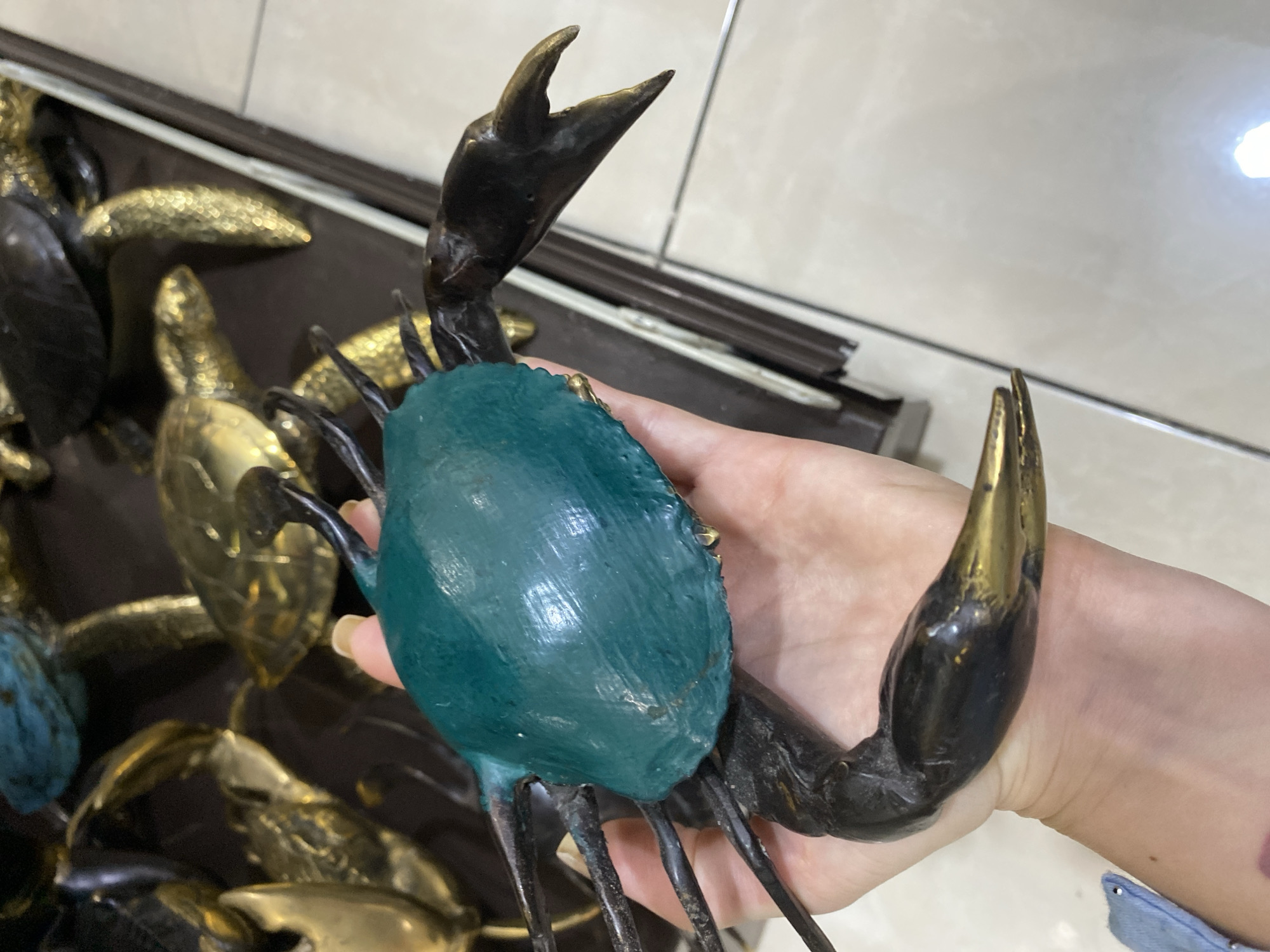 A hand holds a green and black crab statue in front of a display of similar gold statues. The crab statue is black and has golden tips on its claws. The crab's body is a smooth green color. The person holding the crab statue has light skin and a visible blue denim jacket. The background is tile. It is daytime. This photograph was taken indoors, and it may be in a retail setting, likely a souvenir shop. Given the style of items shown, it is possible this image was taken in Nusa Dua, Indonesia, as the prompt states.
