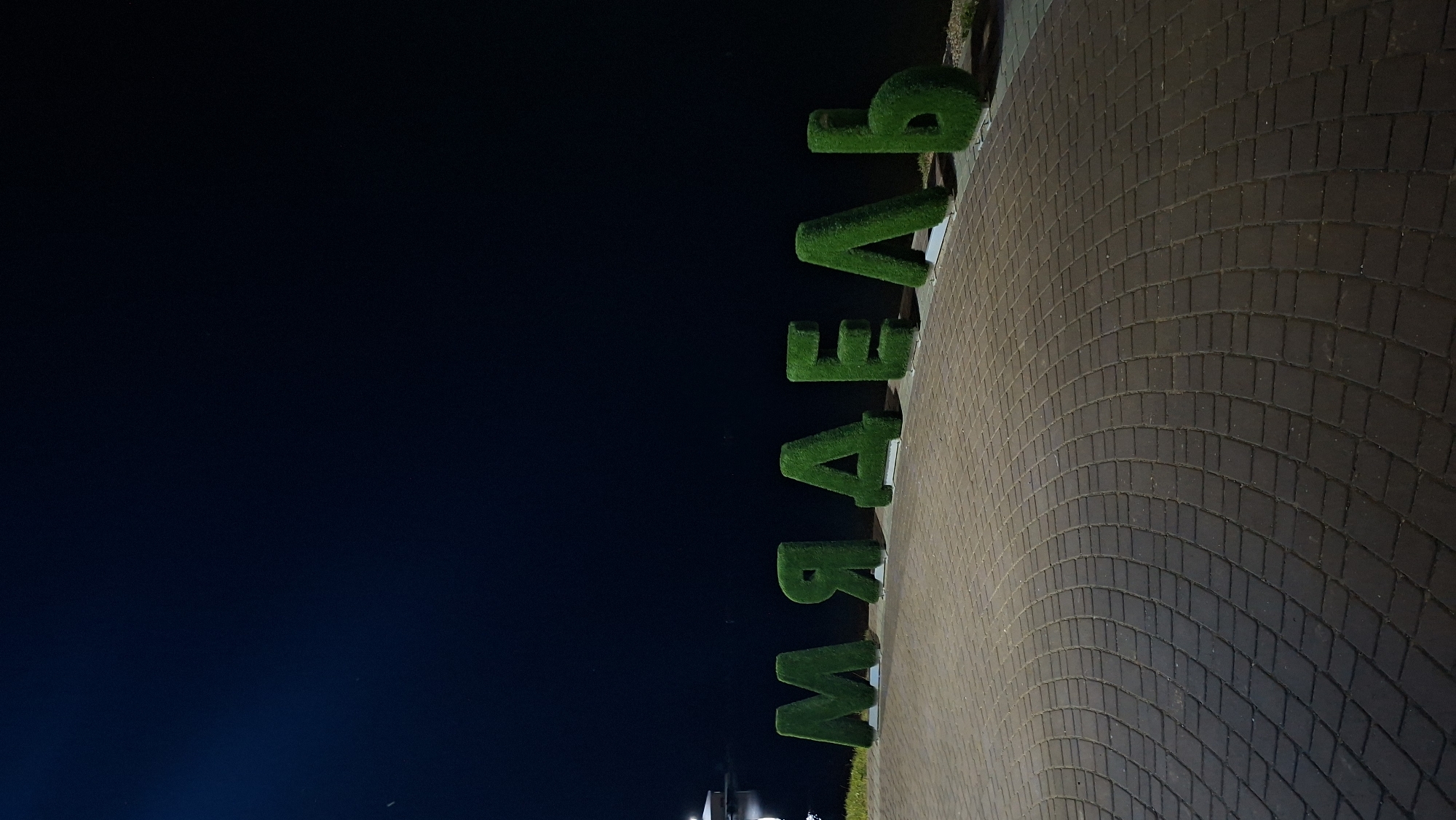 The image shows large, grass-covered letters spelling "МЯДЕЛЬ" (Myadzyel) in Cyrillic script. The letters are arranged in a line along a paved walkway made of dark cobblestones. The walkway curves gently to the right in the foreground. Behind the letters, a dark body of water is visible, with a hint of a distant structure or lights on the far bank. The sky above is a deep, dark blue, indicating it is nighttime. The scene appears to be a waterfront area, likely in the city of Myadzyel, Belarus, given the inscription on the letters. There are no people visible in the image. The lighting suggests artificial light sources illuminating the letters and walkway.