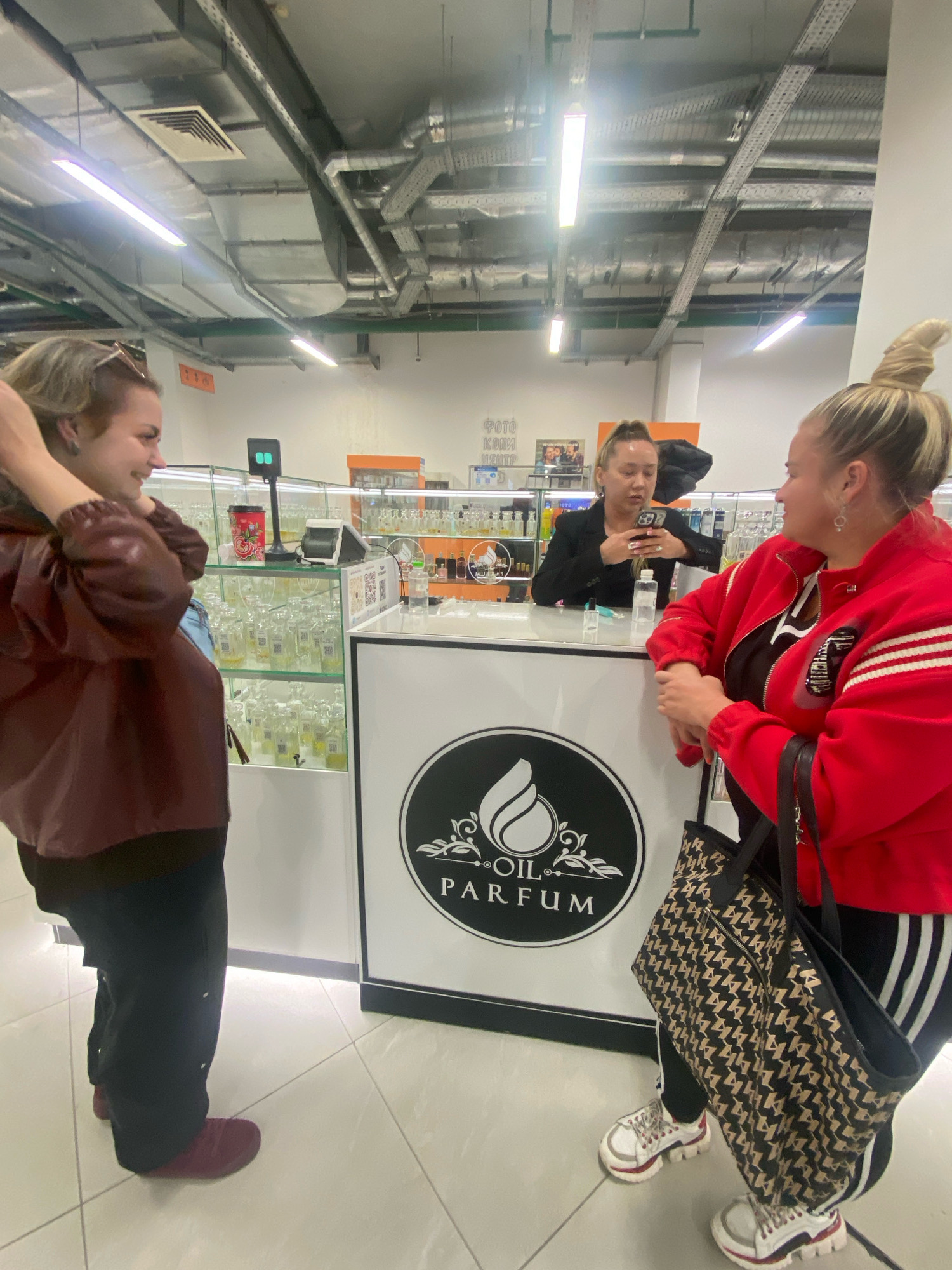 In an indoor commercial setting, likely a retail store in Kazan, Russia, three women are present. The environment appears to be a perfume shop, with rows of glass display cases filled with bottles of various sizes and colors. Overhead, industrial lighting fixtures and exposed ventilation ducts are visible.
In the foreground, a woman on the left, with short blonde hair, is wearing a dark reddish-brown bomber jacket over a darker garment and black pants. She is facing to her right, looking towards another woman. Her left arm is raised to her head, and she is smiling.
In the middle, behind a white counter, a woman with blonde hair pulled back in a ponytail is wearing a black blazer. She is holding a smartphone in her right hand, and her gaze is directed downwards at the device. A clear bottle of liquid and a red disposable cup with a floral design are on the counter in front of her. A small black electronic device with two green lights is also on the counter.
To the right, a woman with blonde hair styled in a high bun is wearing a bright red zip-up jacket with white stripes on the sleeves, over a black and white striped top, and black athletic pants with white stripes. She is looking to her left, engaged in conversation with the woman in the brown jacket. She is holding a large tote bag with a repeating dark brown and beige geometric pattern. She is wearing white athletic shoes with red and black accents.
The counter has a circular logo featuring a flame-like emblem above the text "OIL PARFUM". The floor is tiled with large light-colored square tiles.
