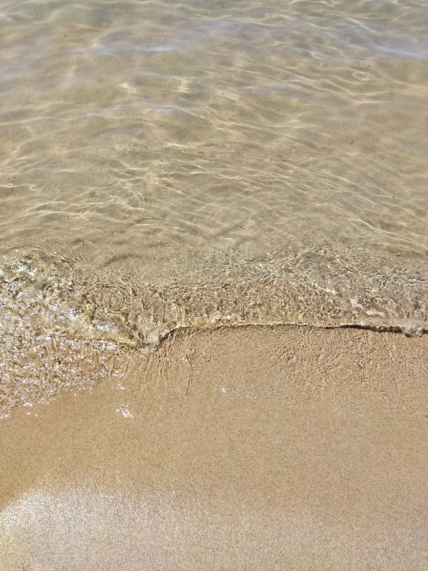 The image shows a close-up of shallow, clear water lapping onto a sandy beach. The water reflects the sunlight, creating shimmering patterns on the sand. The sand itself is fine-grained and a light tan color. The scene is natural and peaceful, with no signs of human activity or urban environment. The location is not discernible from the image itself, and it is impossible to determine if it is in Kara-Oy, Kyrgyzstan based on visual evidence alone.
