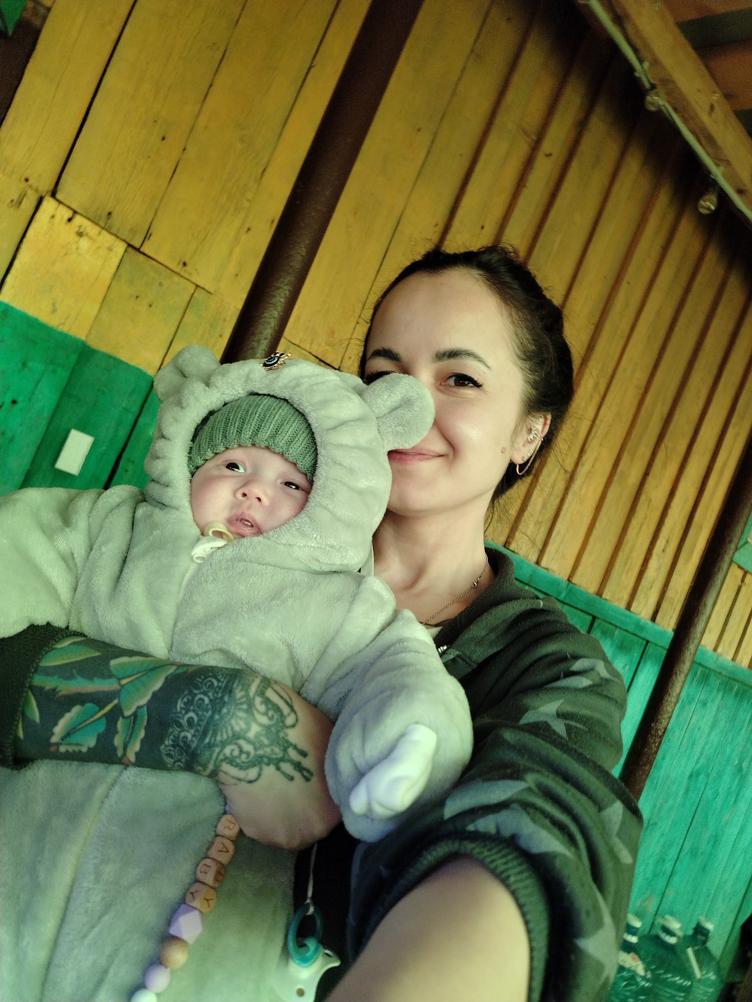A woman holds a baby in her arms. The baby is wearing a gray, hooded outfit with ears and a green knit hat. The woman has dark hair pulled back and is wearing a camouflage-patterned jacket. She has a tattoo on her forearm. The background consists of a wooden structure with green and yellow planks. The wooden beams suggest that the location could be an outdoor covered space like a porch or gazebo. The wooden structure suggests a rural or semi-rural setting, which is consistent with Гожан, Russia being a small village.