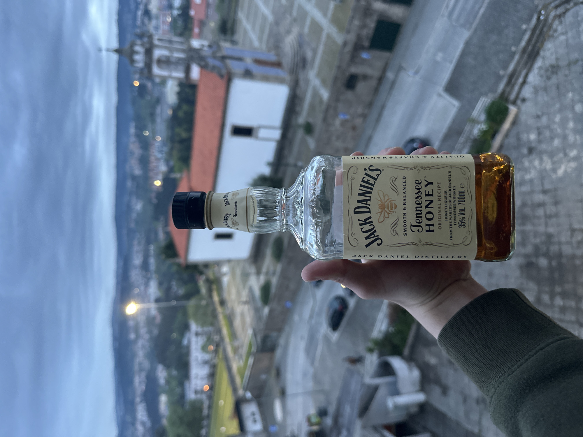 The image depicts a hand holding a bottle of Jack Daniel's Tennessee Honey whiskey against the backdrop of Vila Real, Portugal. The whiskey bottle is predominantly displayed in the foreground. The background features a cityscape with buildings, a church steeple, and a cloudy sky, likely taken from an elevated position. The focus is on the Jack Daniel's bottle, with the city serving as a secondary element to give the photo context. The hand holding the bottle appears to be wearing a green sleeve.