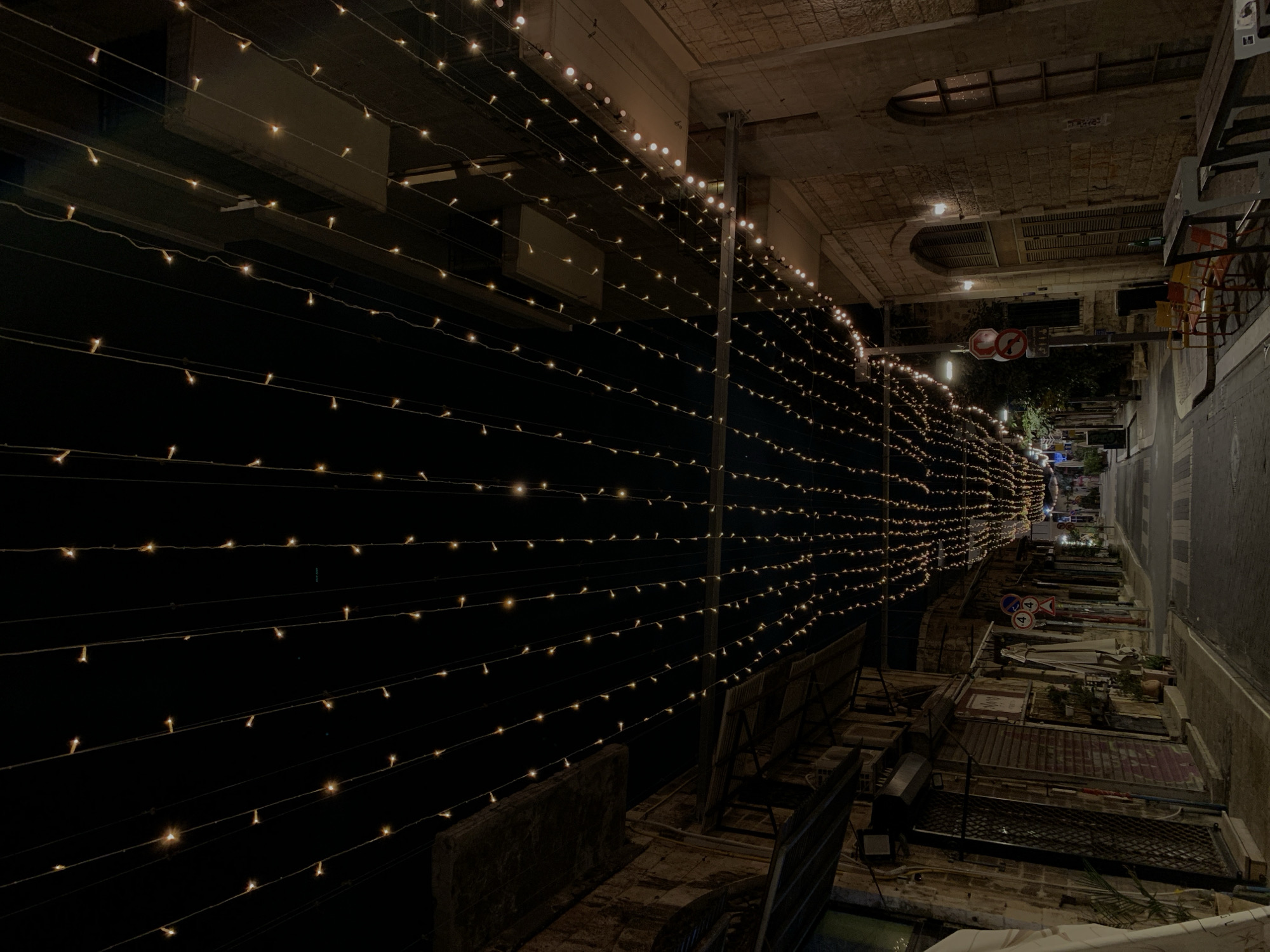 The image depicts a narrow street in Tel Aviv, Israel, illuminated by strings of lights spanning overhead. The scene appears to be at night. The strings of lights create a tunnel effect, drawing the eye down the street. The architecture is characterized by aged stone buildings with arches and windows above. The street itself appears clean and well-maintained, with various objects like chairs and tables suggesting a dining or gathering space. The street continues away in the distance, illuminated by streetlights. There are no people visible.