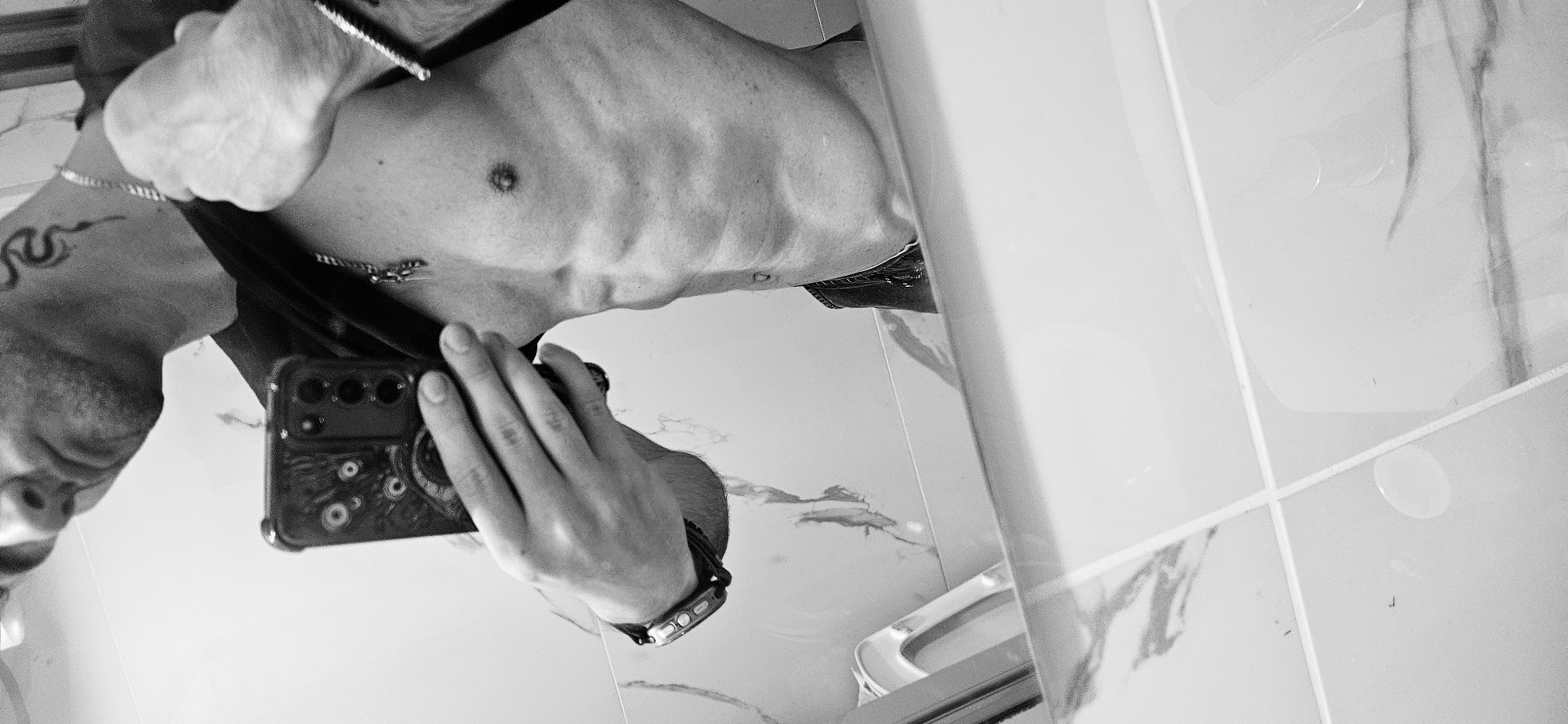A shirtless male takes a selfie in a tiled bathroom. The man has a snake tattoo on his upper left arm and wears jewelry around his neck and wrist. He holds a phone in his right hand. A toilet is visible in the background. The location is indeterminable from the image but presumed in Plovdiv, Bulgaria.