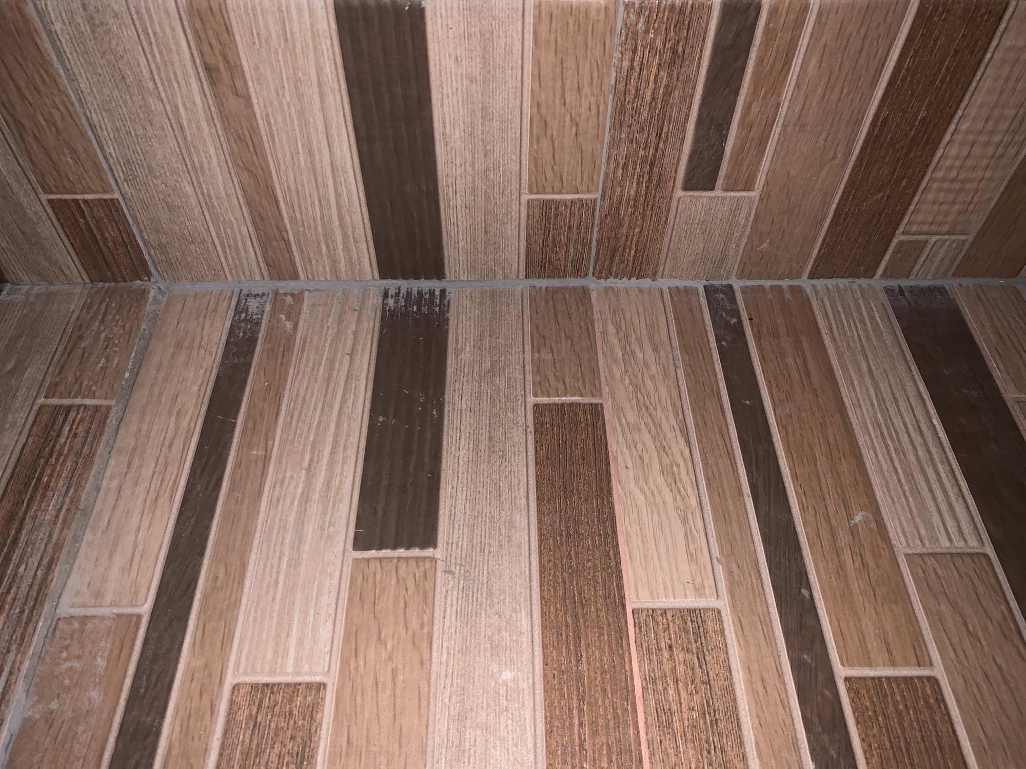 The image shows a tiled floor and lower wall section, with a wood-grain pattern. The tiles are primarily arranged in long, narrow strips, with alternating light and dark brown shades mimicking wood. Some shorter tiles are interspersed among the longer ones, creating a staggered effect. The grout lines are a light gray color. The floor meets the wall at a distinct line, where the same tiles continue up the wall. The floor appears slightly dirty in places. The exact location in Santa Ana, Venezuela, cannot be determined from the image alone. There are no people, subjects, objects, actions, or interactions depicted.