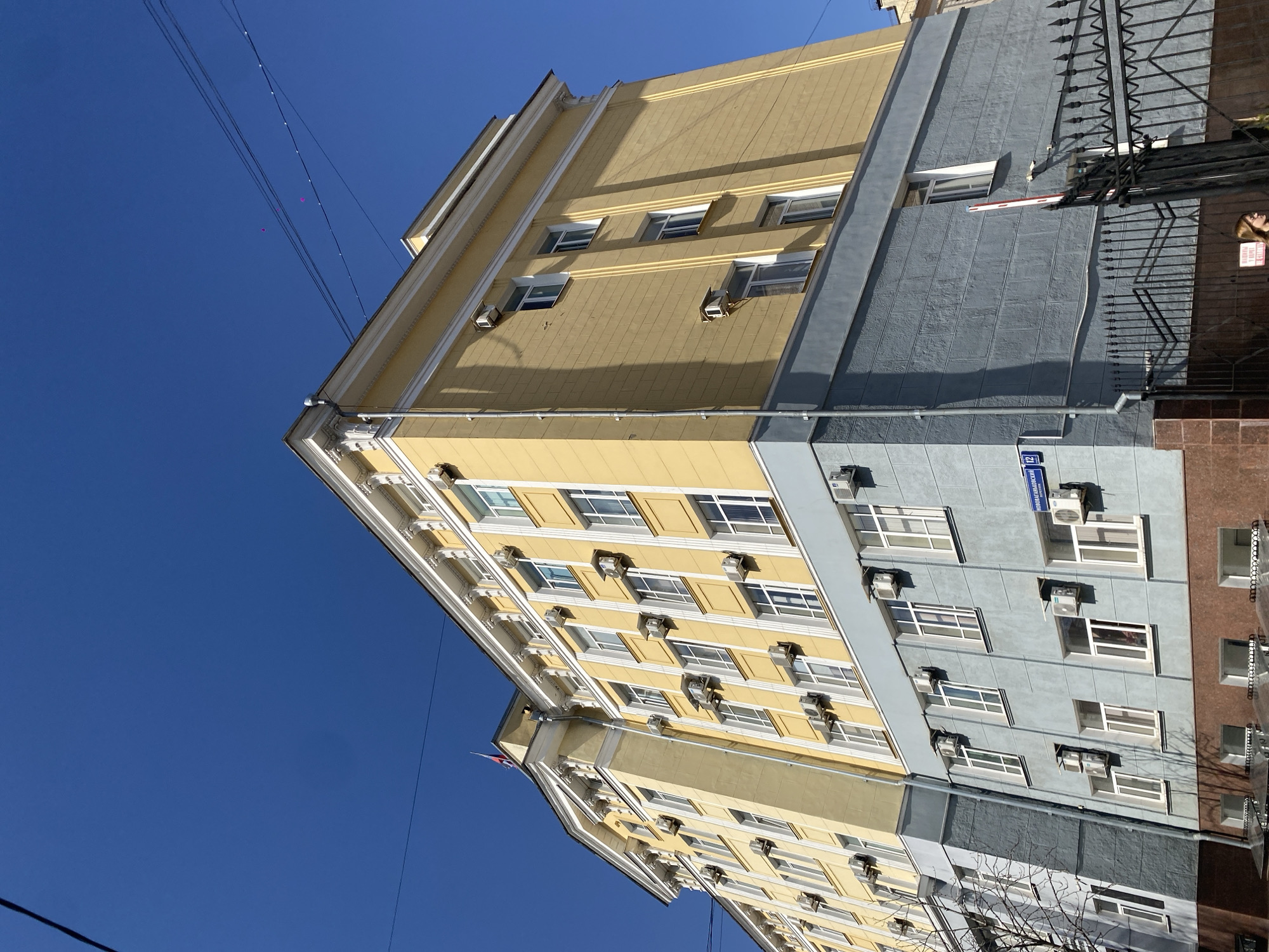 The image depicts a multi-story building in Moscow, Russia. The building has two distinct sections, one painted yellow and the other gray. Both sections have windows with air conditioning units installed. A street sign reading "Novozavodskoy Pereulok 12" is visible on the gray section. The sky is clear and blue. Wire lines are also visible across the sky. A black metal fence is also visible on the right. The building appears to be residential.