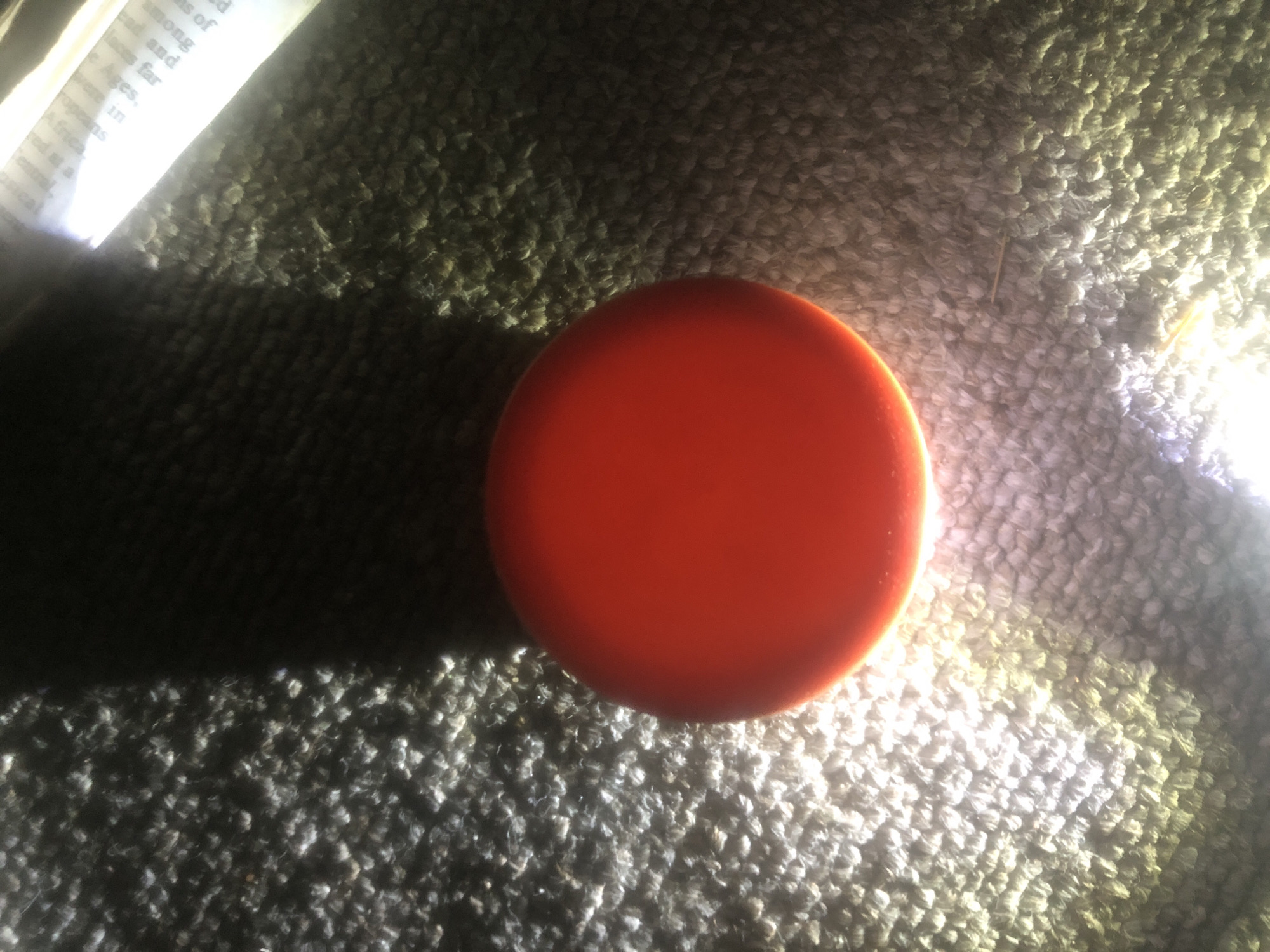 A red, spherical object is centered on a textured, gray surface, possibly carpet or a similarly soft material. The object is brightly lit from below, causing it to glow and cast a shadow upwards and to the left. To the right of the red object, a portion of a white paper with text is visible, also illuminated by light. The overall scene is dimly lit, with the primary light source appearing to be from below the red object. There is no indication of a specific location in Jalingo, Nigeria, as the image only shows a close-up of the object and its immediate surroundings.
