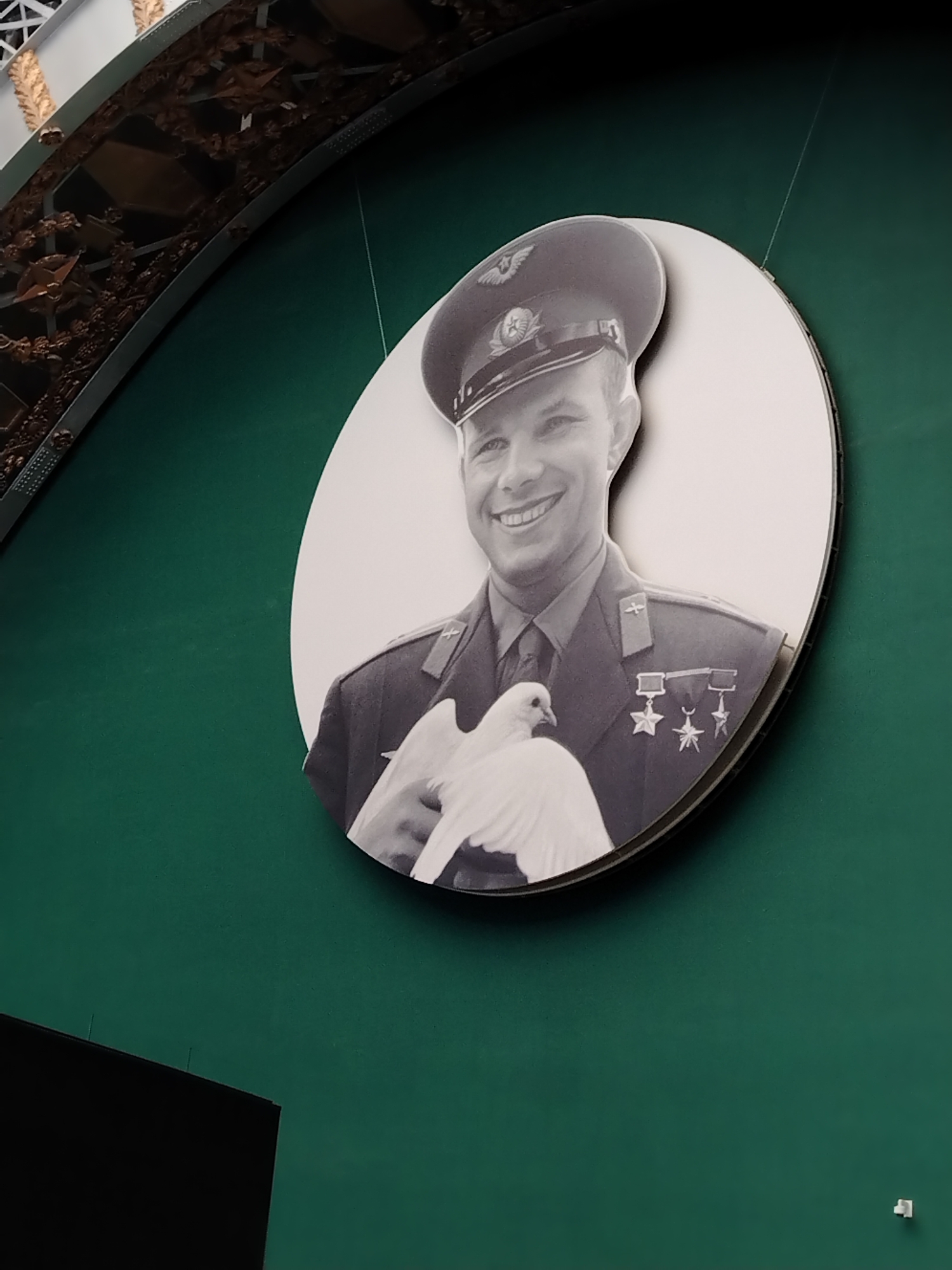 The image shows a black and white circular cutout of Yuri Gagarin, the first human in space, holding a white dove. He is wearing a military uniform and a cap with a star insignia. The cutout is mounted on a dark green wall. Above the cutout, a portion of an arched structure with ornate bronze decorative elements is visible. This scene is located at the Cosmonautics Memorial Museum in Moscow, Russia.