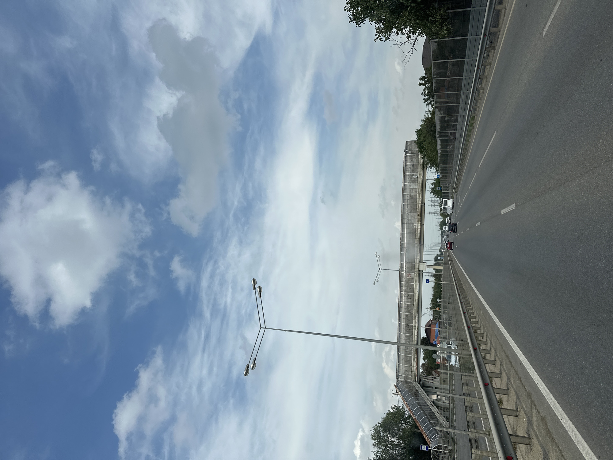 A perspective shot from a moving vehicle on a highway leads the viewer towards a pedestrian overpass and distant buildings under a partly cloudy blue sky. The highway has multiple lanes and is bordered by metal guardrails and sound barriers. On the left side, a pedestrian overpass with a glass enclosure arches over the road, with a ramp leading up to it. Streetlights are visible, one prominently in the foreground. Several cars and a bus are driving on the highway. The environment appears to be on the outskirts of a city, possibly Beryozovy, Russia, given the architecture and vegetation.