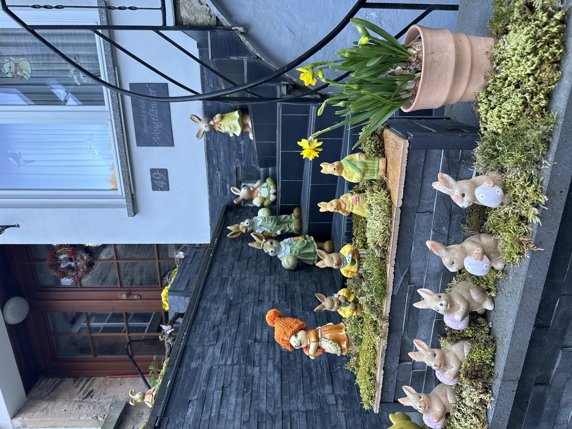 The image shows a set of stone steps decorated with Easter decorations leading up to a house in Beilstein, Germany. There are multiple ceramic bunny figurines with painted details arranged on the steps. Some are holding eggs. One figurine is a doll wearing an orange hat and brown dress. The steps are partially covered with moss and have a flower pot with yellow flowers on the top step. The house is visible in the background, with a window and the number "49" next to a sign reading "Monika & Rolf Voigtländer". A wreath is hanging on the wooden front door. A black metal railing lines the staircase. The environment is likely residential.