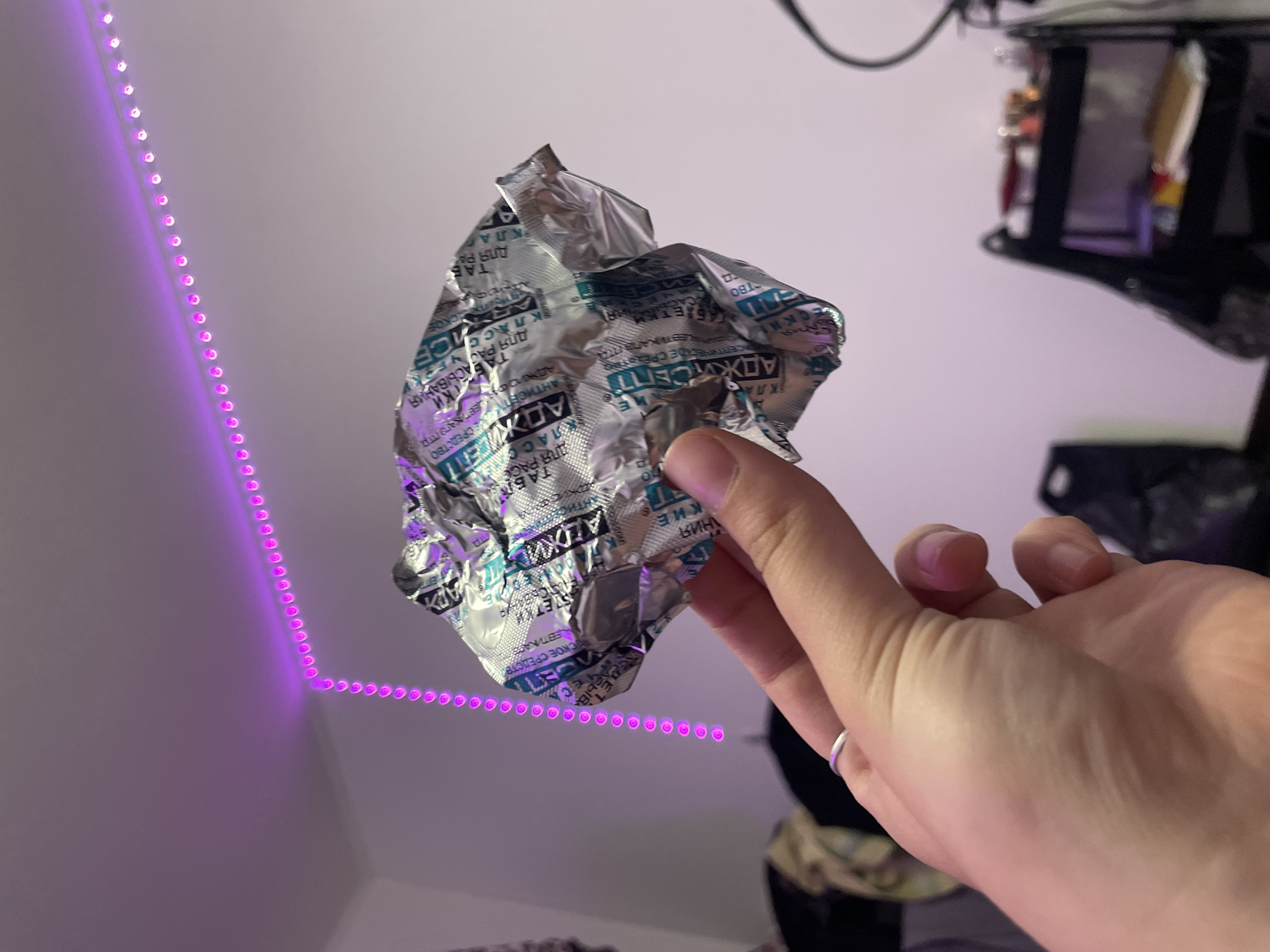 A crumpled silver foil blister pack, likely containing medication, is held in a person's left hand, which has a silver ring on the ring finger. The blister pack has text in a blue and white font. The background is a room with a white wall. A string of purple LED lights is mounted along the top of the wall, creating a border. To the right, a black shelf unit with various items is visible. The image is taken in a bedroom. The location of Vaganovo, Russia is irrelevant to the image content.