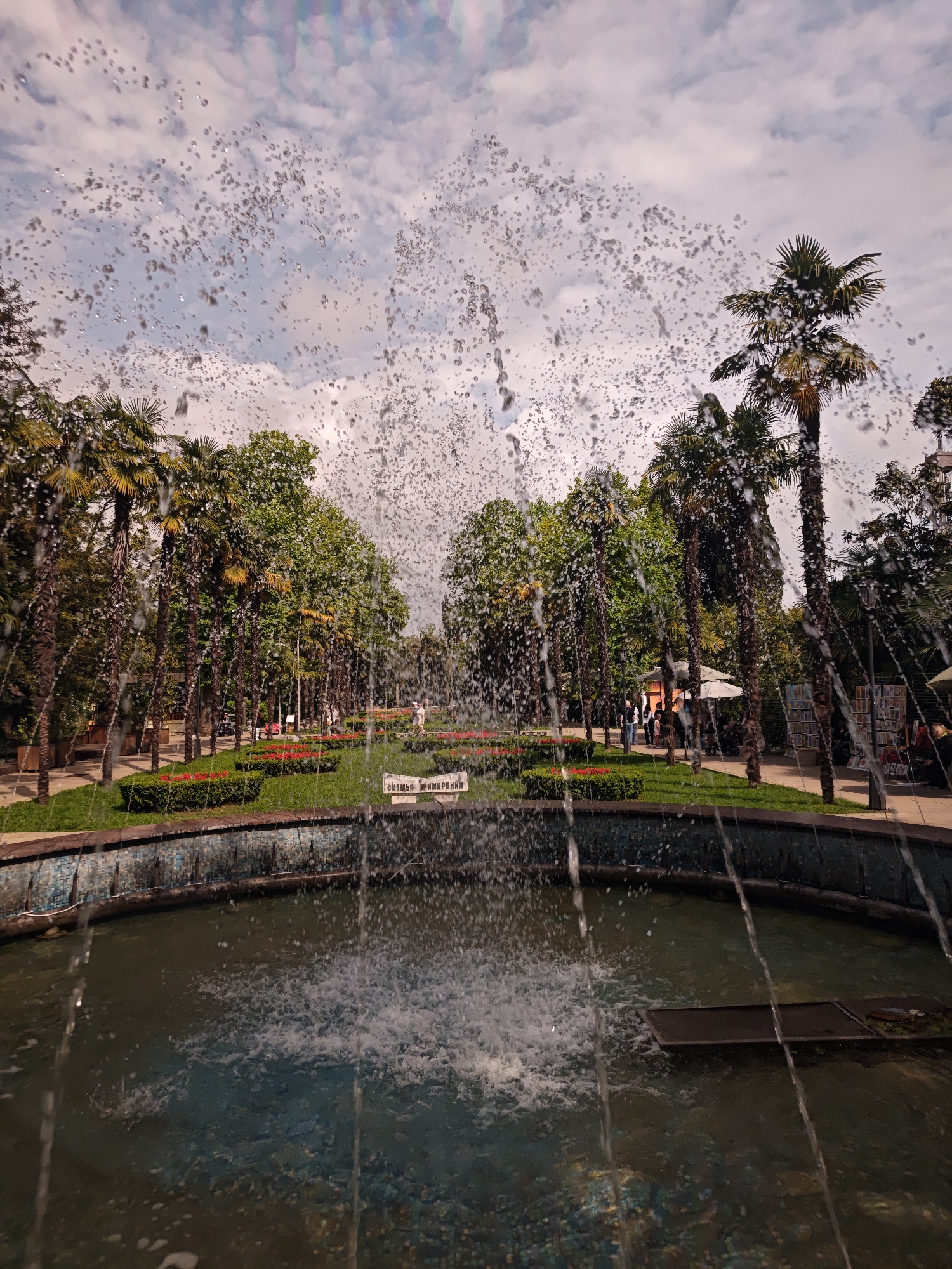 The image depicts a fountain in a park in Sochi, Russia. The fountain's water is spraying upwards and outwards, partially obscuring the view. Behind the fountain, there is a well-manicured park with tall palm trees lining a path. There are flowerbeds with red flowers and trimmed bushes. People can be seen walking in the background. The sky is cloudy. The water in the fountain's pool is dark.