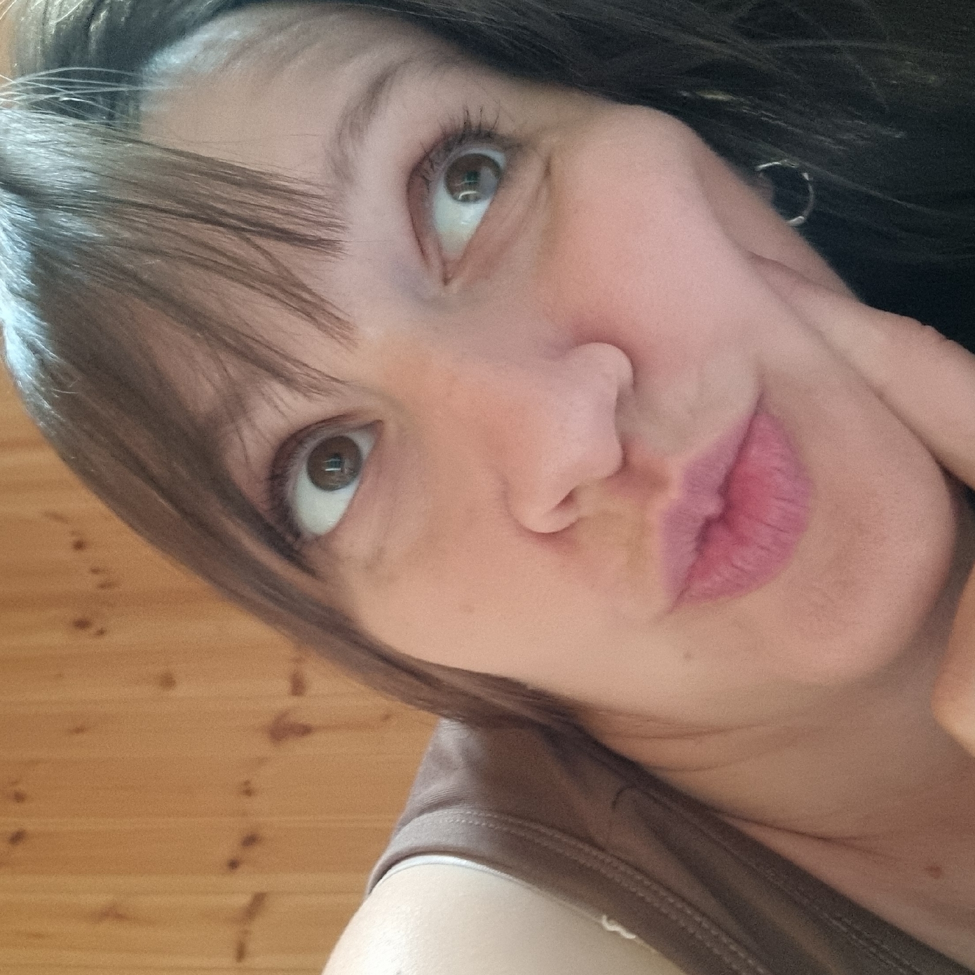 The image is a close-up, low-angle selfie of a woman with brown hair and brown eyes. She is wearing a brown tank top. Her face is tilted upwards and to the right, with her left hand resting on her cheek and her mouth slightly pursed. Her right eye is looking directly at the camera, while her left eye is looking upwards and to the right. The background appears to be a wooden wall with horizontal planks. The lighting is warm and natural. The specific location in Dunnae-myeon, South Korea is not discernible from the image.