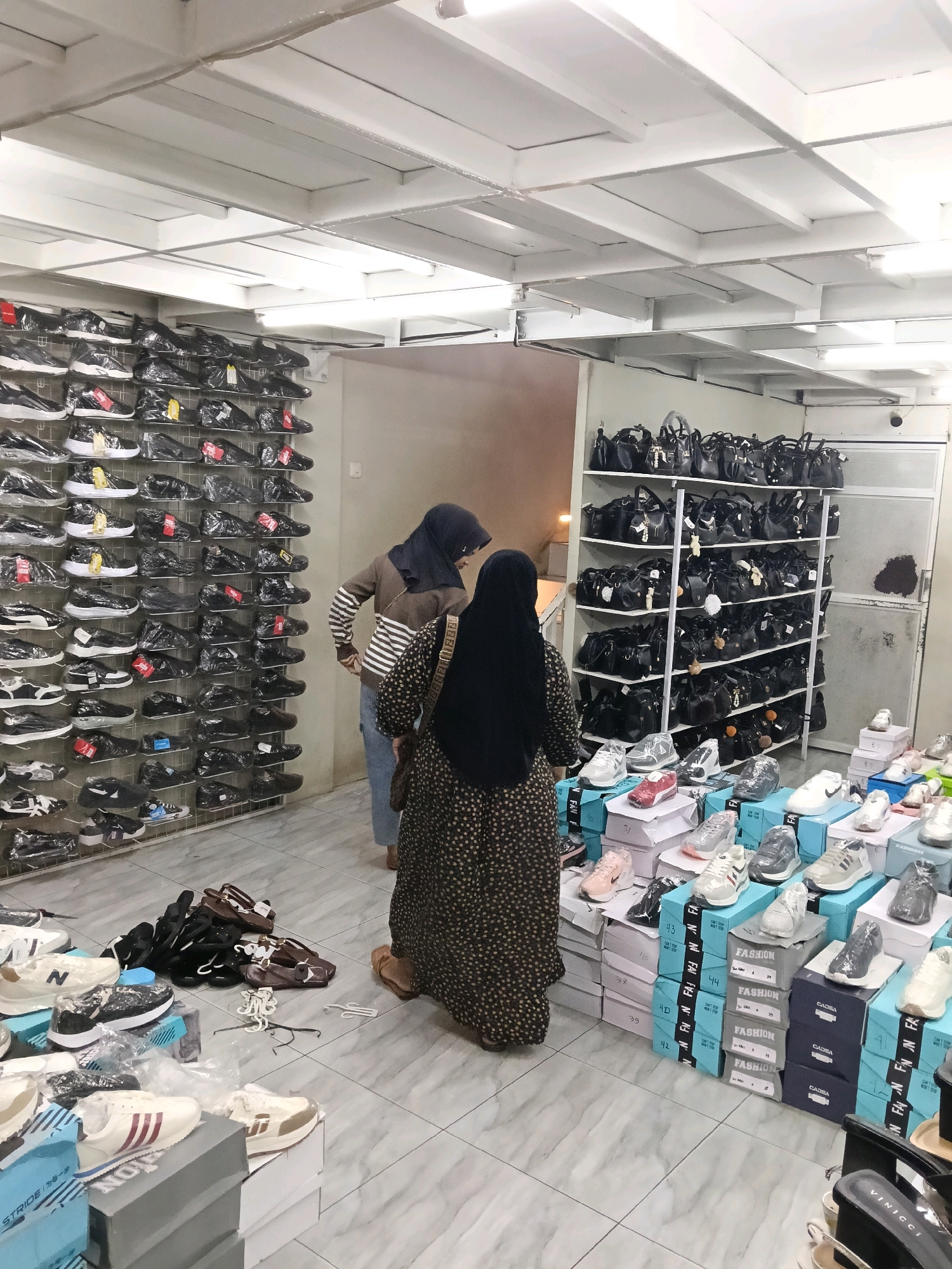 The image depicts the interior of a shoe store, likely located in Dengok, Indonesia. The store is filled with various footwear displayed on shelves and stacked on the floor. Two women are present in the store. One woman, wearing a headscarf and a striped sweater, is examining shoes on a wall display. The other woman, also wearing a headscarf and a patterned dress, is standing near a display of shoes on the floor. The store has a tiled floor and a white ceiling with exposed beams. Shelves on the right side of the image are stocked with black handbags. Numerous shoe boxes are visible, stacked on the floor, some labeled with sizes and the word "FASHION." Several pairs of shoes are also displayed on the floor. The overall impression is of a bustling retail environment with a wide selection of footwear.