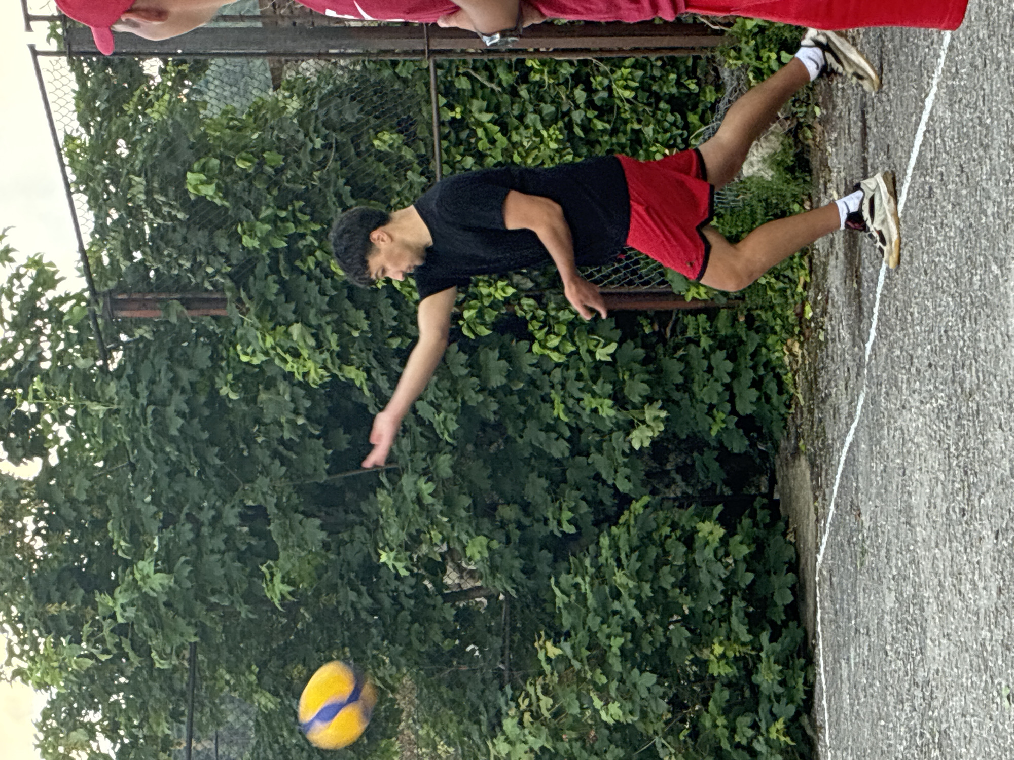 The image depicts an outdoor scene in Klisura, Bulgaria, where two males are engaged in a volleyball activity on a textured, dark gray asphalt court.
In the center-left, a male with short, dark hair, wearing a black short-sleeved t-shirt, red athletic shorts, white socks, and light-colored sneakers, is captured in motion. His right arm is extended forward and slightly upward, indicating he has just struck or is about to strike a yellow and blue volleyball, which is visible in mid-air to his upper left, showing slight motion blur. His body is angled, with his left foot forward and right leg extending backward, suggesting a follow-through or preparatory stance.
On the far right, a second male is partially visible, from his upper torso upwards. He wears a red baseball cap and a red t-shirt, with a silver-colored watch on his left wrist. He appears to be observing the action, facing towards the central player.
The background consists of a weathered metal chain-link fence running horizontally across the mid-ground, behind which dense, green deciduous trees and bushes form a lush backdrop. A bright, possibly overcast, sky is visible above the foliage. A distinct white line, likely a court boundary, is painted on the asphalt court in the foreground.
