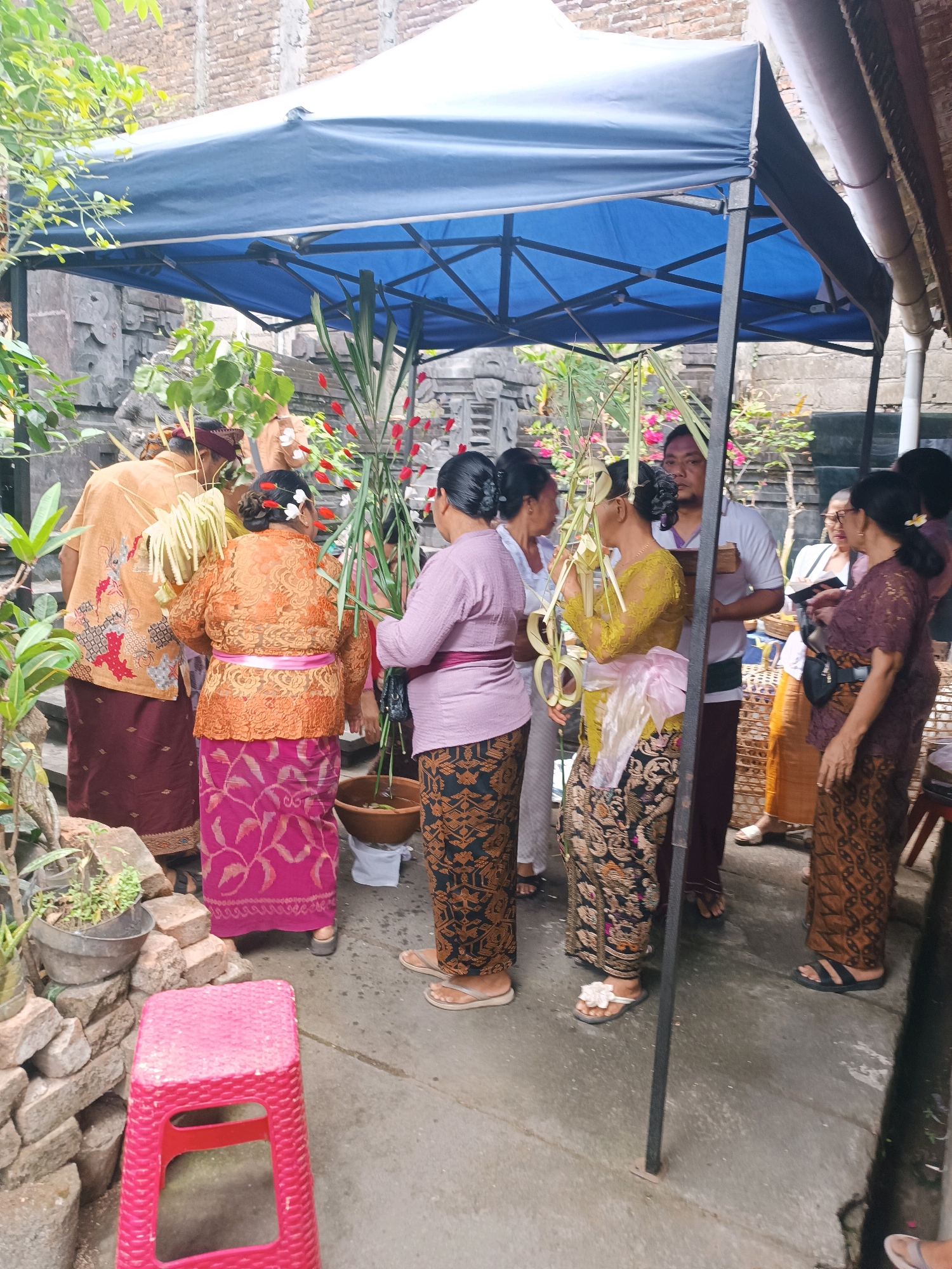 The image shows a group of people gathered under a blue canopy in what appears to be an outdoor setting with traditional Balinese architecture in the background. The people are dressed in traditional Indonesian clothing, with several women wearing intricately patterned sarongs and lace tops. Some are holding decorative items made from palm leaves. The ground is paved, and there are potted plants and stone structures visible. The scene suggests a cultural or religious gathering. The overall impression is one of community and tradition. Based on the context provided, the location is identified as the city of Tabanan, Indonesia.