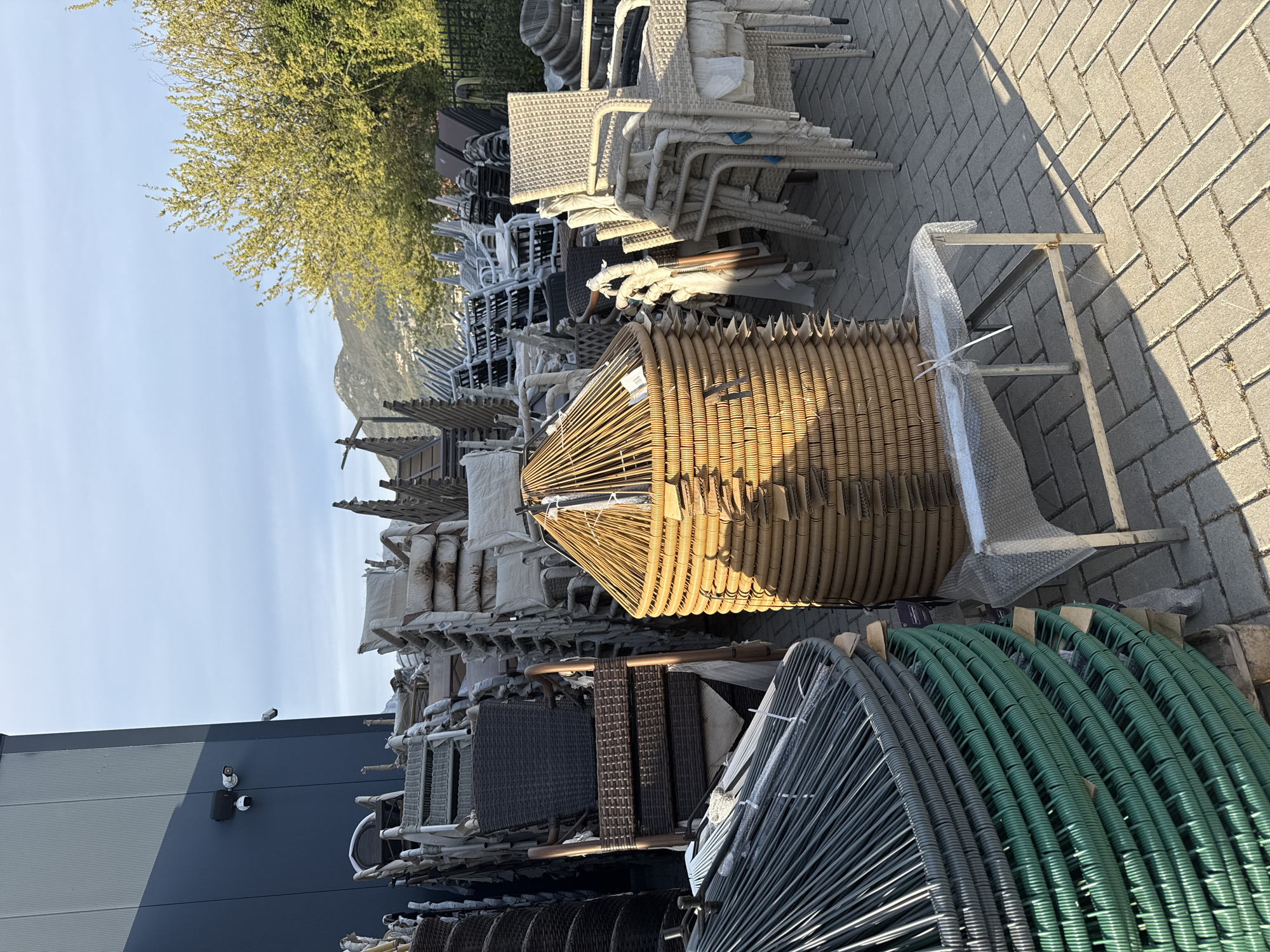 The image shows a large collection of outdoor furniture stacked outdoors, likely for storage or display. The furniture includes wicker chairs, stacked dining chairs, and possibly lounge chairs. The scene is located outdoors, with a gray building visible on the left side and a paved area with brick patterns on the right. There is a tree in the background and a mountain in the distance, suggesting the furniture is stored near a retail location in an area with natural landscape. The specific city or location is not identified.
