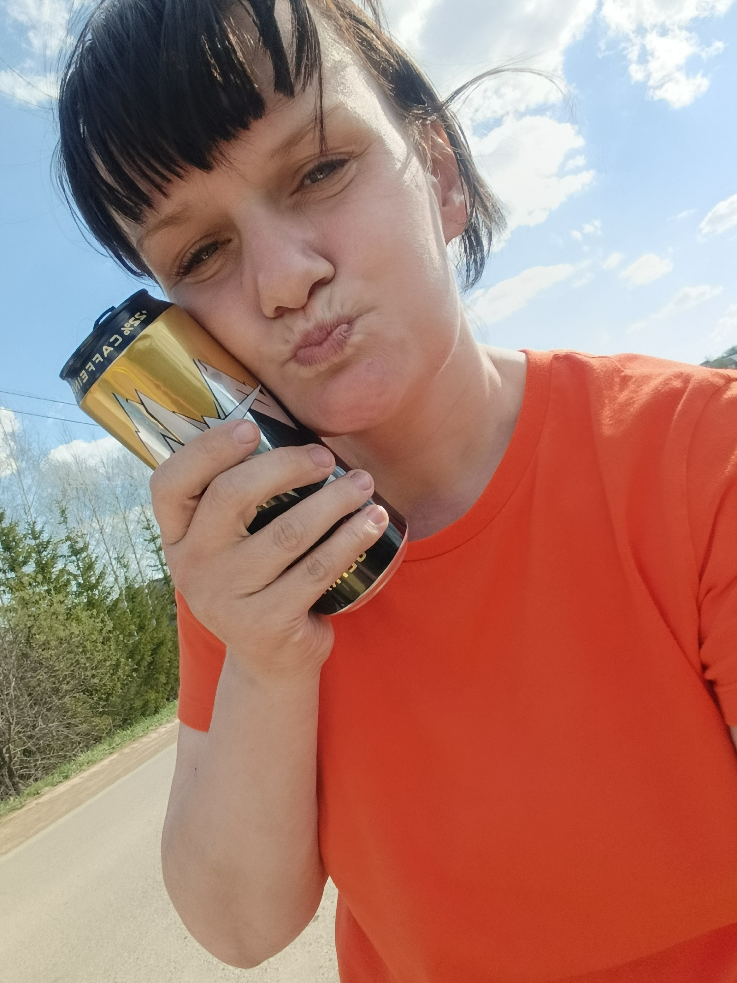 A selfie shows a woman with short dark hair and bangs, wearing an orange t-shirt. She is holding a gold and black can against her cheek and making a kissing face. The can reads "35% CAFFEINE." The background includes a blue sky with scattered clouds, green trees, and a road. The location appears to be outdoors, possibly in a rural area within the city of Фоки, Russia.