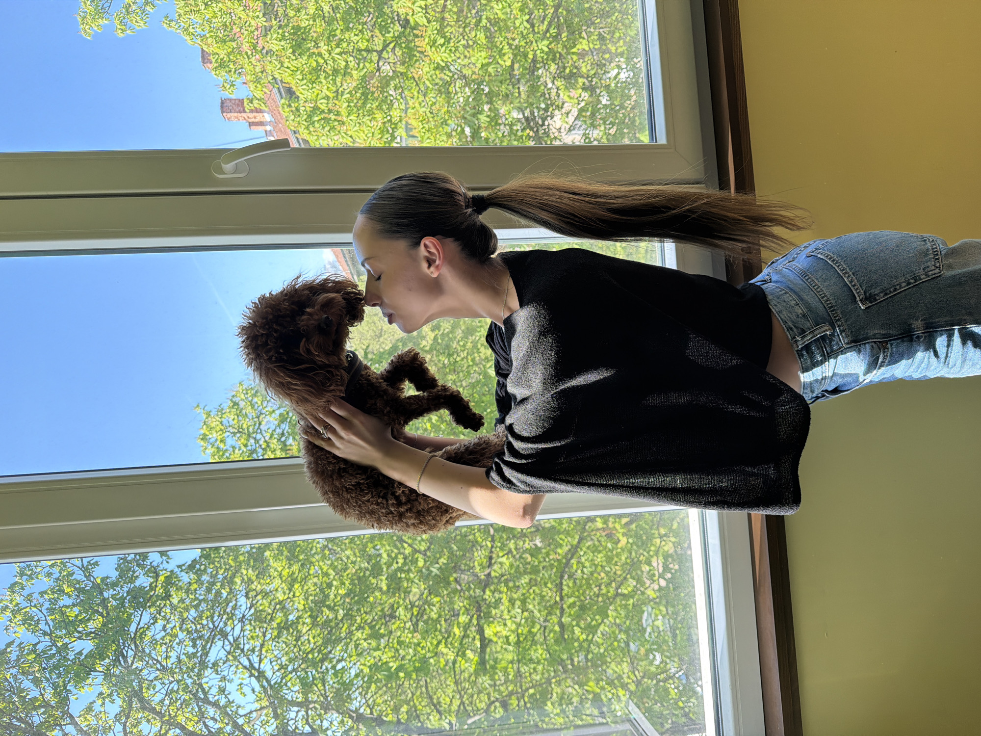 A woman with a high ponytail is standing indoors by a large window, holding a brown poodle. She is wearing a black long-sleeved shirt and blue jeans. The window shows a view of a building and trees under a blue sky. The woman and the dog are positioned close to each other, their faces nearly touching. The wall near the window appears to be light yellow. Without additional information or details within the image that definitively places it in Sofia, Bulgaria, it's impossible to determine the location.