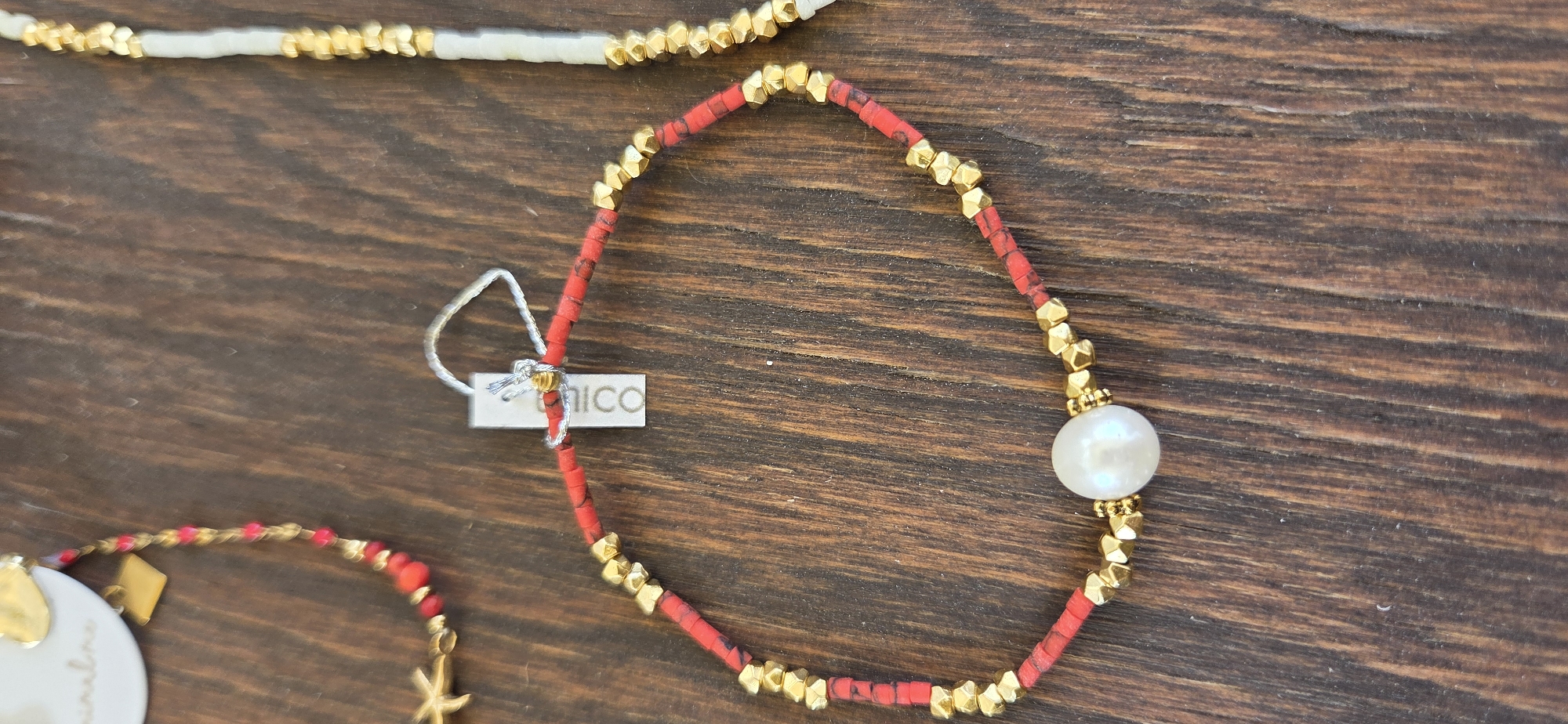 A close-up, overhead shot shows three beaded bracelets arranged on a dark wooden surface. The bracelet in the foreground is made of red beads interspersed with gold, hexagonal beads, and features a single large white pearl at its center. A white tag with blue text is attached to this bracelet with a silver string. Another bracelet, to the left and slightly out of focus, is red and gold with a gold starfish charm. A third bracelet, to the right and also out of focus, consists of white and gold beads. The lighting is even and highlights the textures of the beads and the wood grain. No people are visible, and the setting does not provide any specific information about the city of Karlsruhe, Germany.