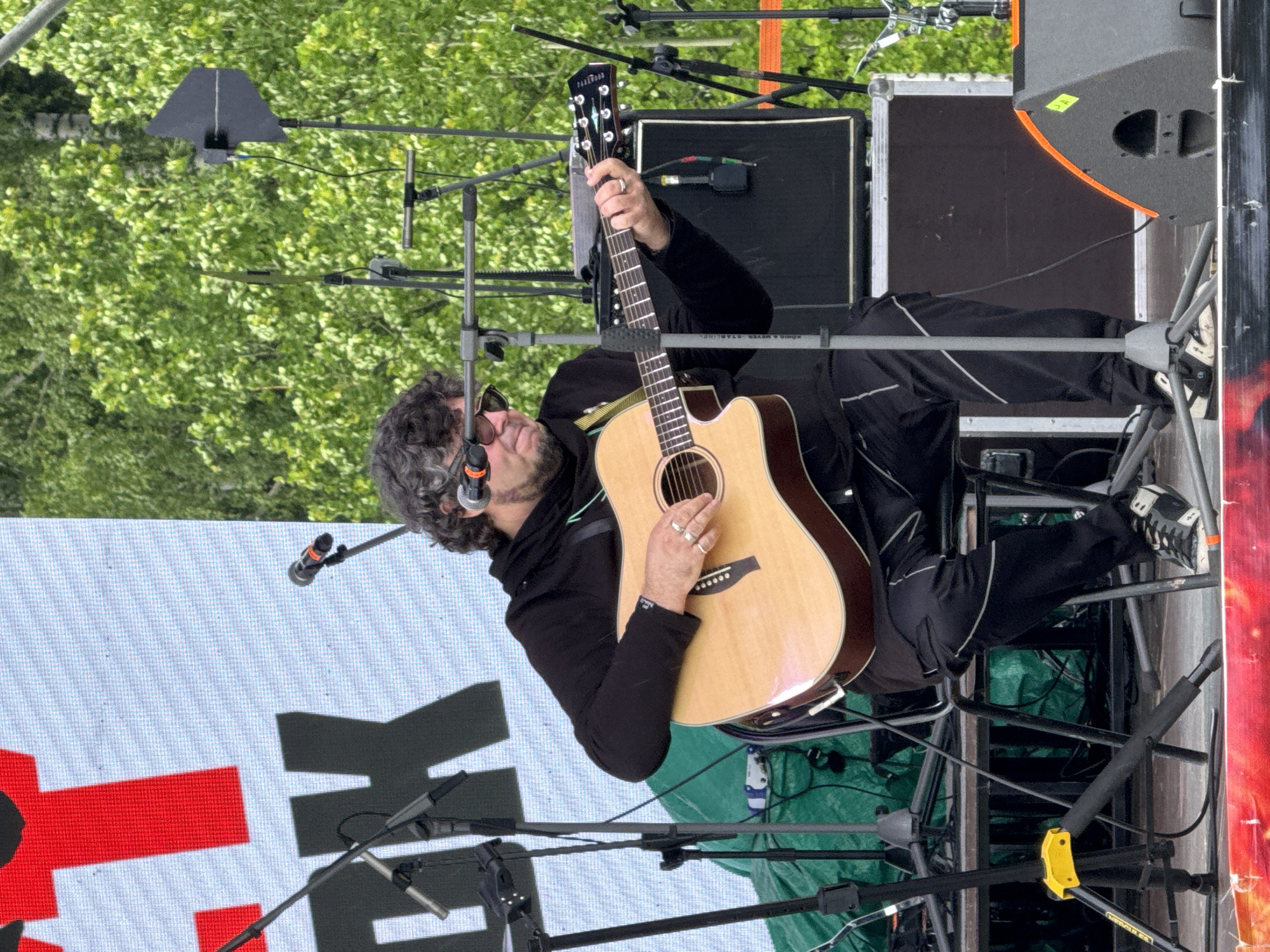 A male musician performs on an outdoor stage during the day. He has curly grey hair, a beard, and wears sunglasses, a dark hoodie, and dark track pants with white stripes. He is playing a light-colored acoustic guitar, with "Fenderwood" visible on its headstock. Two rings are worn on his left hand.
The stage is equipped with microphones on stands, sound equipment including speakers, and metal rigging. A microphone is positioned in front of the musician, indicating he is likely singing while playing. In the background, green trees are visible, suggesting an outdoor setting. A large LED screen or backdrop behind the performer displays partial text in red and black, including a prominent red "K" and another black letter.
Additional visible details include microphone stands, one bearing the text "König & Meyer STARLINE," and a case marked "DIRBOX." A green tarp is visible beneath parts of the stage structure. The scene depicts a live musical performance in Большая Кивара, Russia.