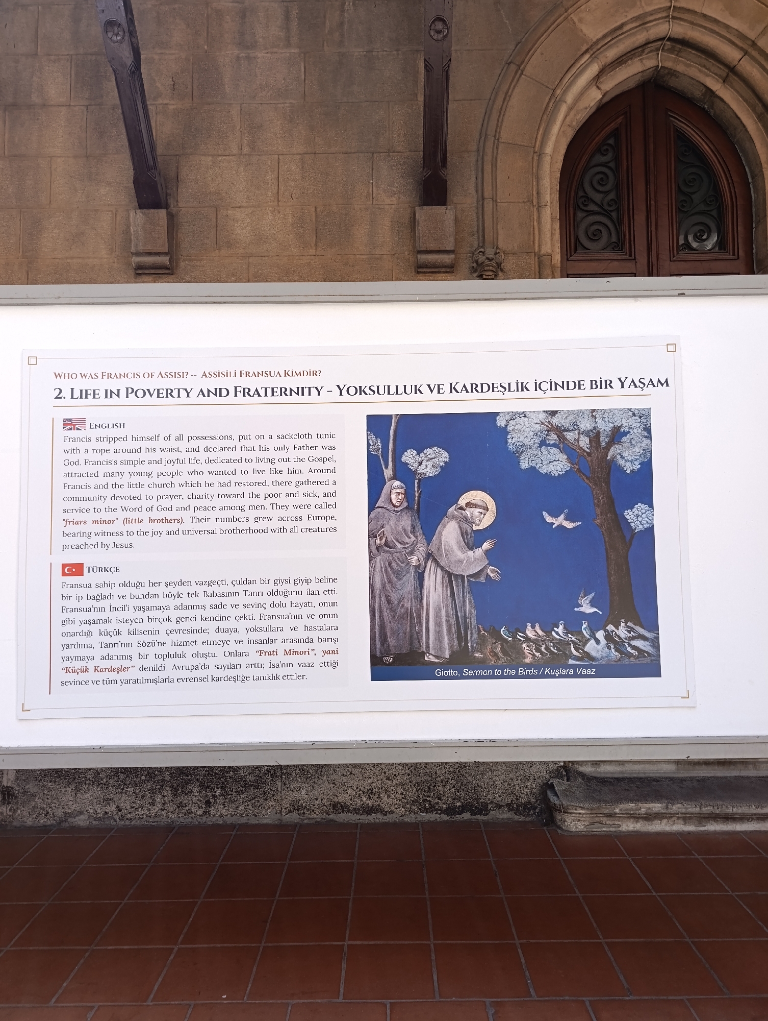 The image shows a framed exhibit with text in English and Turkish and a painting. The exhibit is located on a wall, possibly an exterior wall of a building, given the brickwork visible above the exhibit and the tiled floor below. The painting depicts Saint Francis of Assisi in a brown robe, with a halo, preaching to a flock of birds gathered at the base of a tree. Another friar stands beside him, also in a brown robe. The painting is signed "Giotto, Sermon to the Birds / Kuşlara Vaaz." The text above the painting describes "Life in Poverty and Fraternity" and provides biographical information about Francis of Assisi in both English and Turkish. The setting appears to be an outdoor exhibition space, with an arched doorway visible in the upper portion of the image, suggesting a historical or religious context. The exact location in Beyoğlu, Turkey is not explicitly stated, but the architectural elements and the presence of Turkish text indicate it is within Turkey.