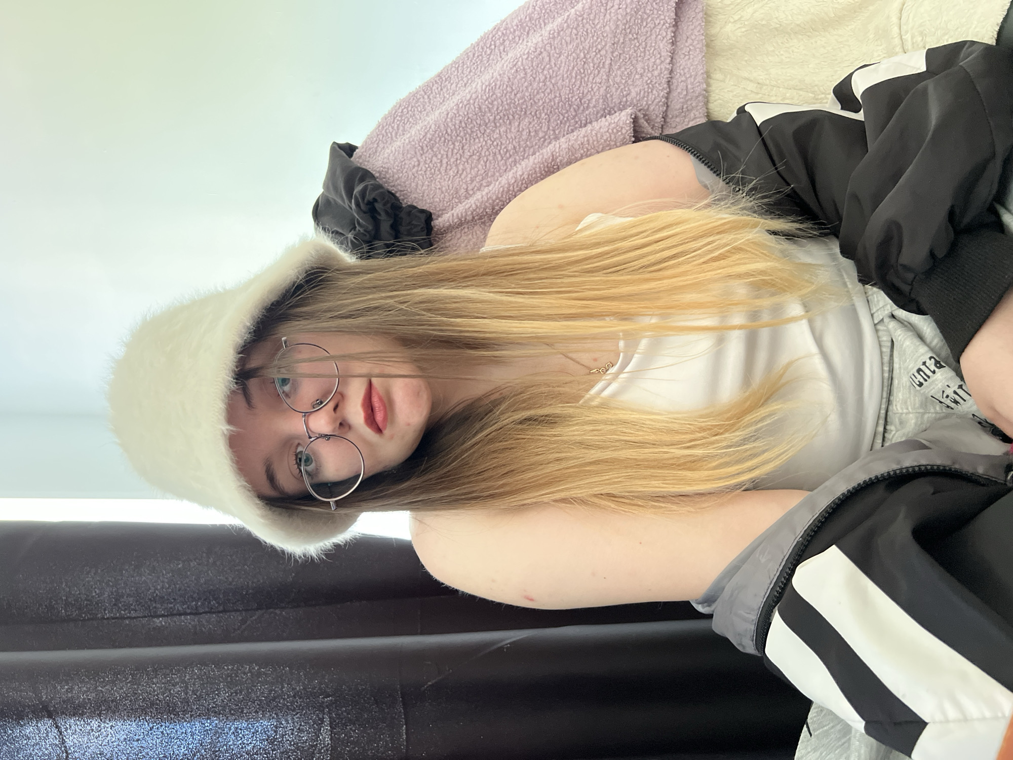 A young woman is lying on a black surface, likely a sofa or bed, wearing a white tank top, a furry white hat, and round glasses. She has long blonde hair and fair skin. She is partially covered by a black and white jacket. A lavender blanket is behind her. The location is not discernible from the image. Pushchino, Russia is not discernible from the image.