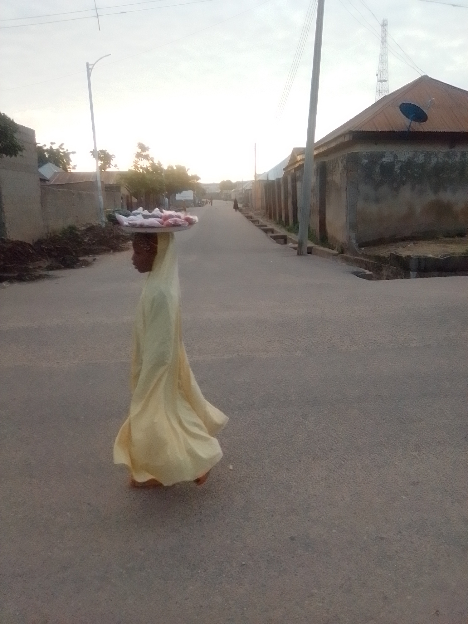 A child is walking down a paved street in Liji Hill, Nigeria, carrying a large tray of raw meat on their head. The child is wearing a light yellow, flowing garment that covers them from head to toe, with only their face and feet visible. The street is lined with buildings on both sides, some with corrugated metal roofs and others with concrete walls. There is a street lamp on the left side of the road. The sky is overcast with a hint of sunlight breaking through, suggesting it is either early morning or late afternoon. The overall scene depicts a common sight in many Nigerian communities where children often assist with family businesses.