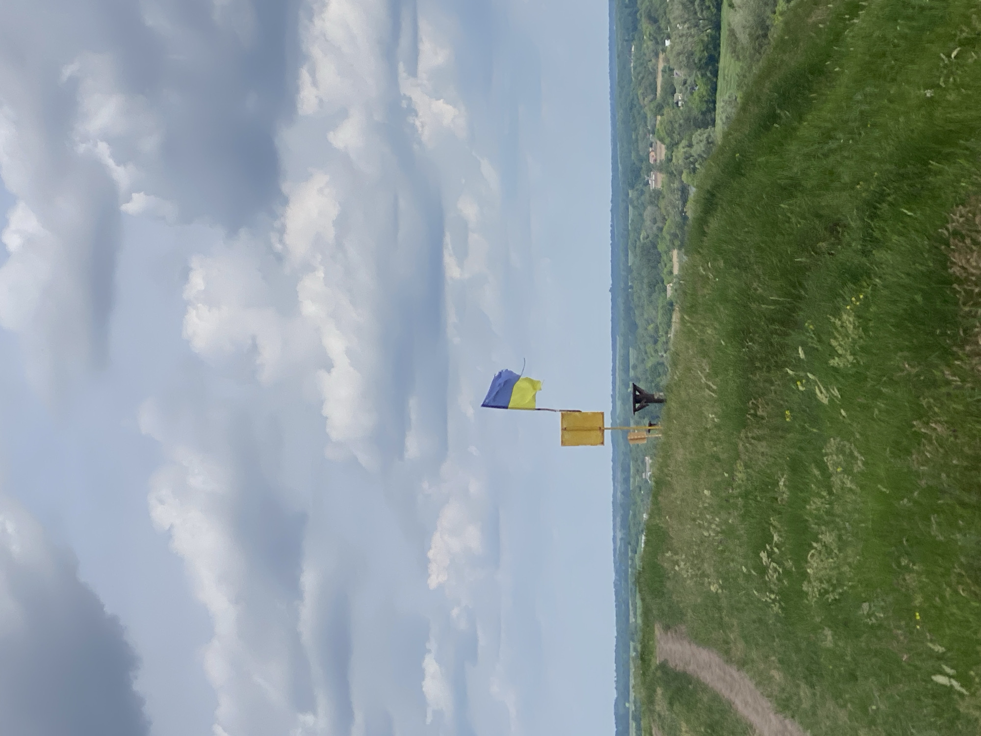 A long shot depicts the Ukrainian flag waving in the wind near a grassy hill in Ustyvytsia, Ukraine. The sky takes up the left half of the frame and is filled with light, puffy clouds. Below the sky, the Ukrainian flag flies from a yellow pole. To the right of the flagpole, the grassy hill rises up and fills the right half of the frame. In the distance, a green treeline and several buildings can be seen. A path leads up the hill from the foreground.