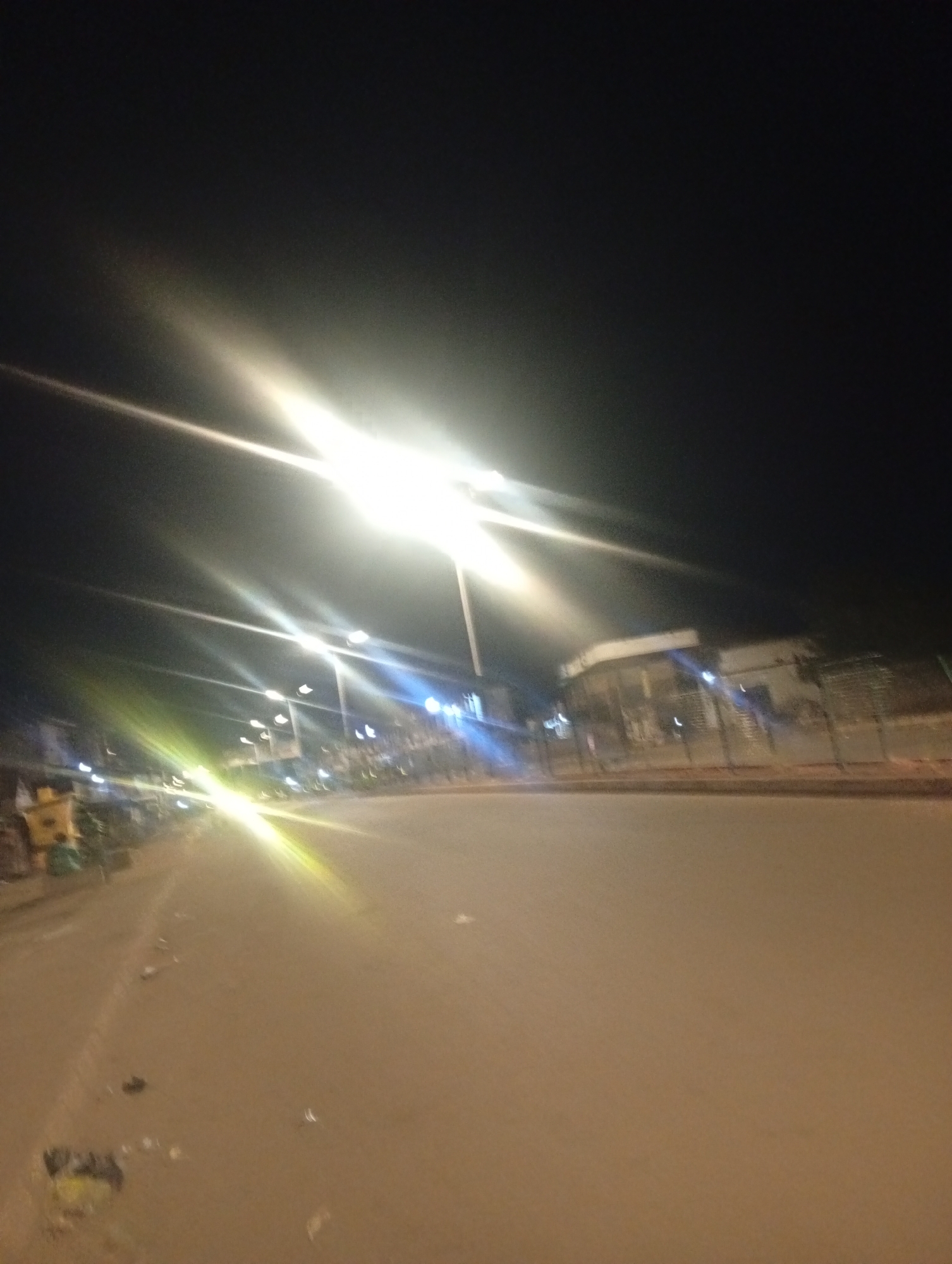 The image depicts a street scene at night in Bukuru, Nigeria. Bright streetlights illuminate the road, creating strong light flares and streaks. The street appears to be paved with asphalt. To the left, there are some objects on the side of the road, including what appears to be a yellow bin or container and some trash. In the background, there are buildings with lights on, suggesting commercial or residential activity. A fence is visible in front of one of the buildings. The overall impression is of a busy street that is well-lit despite the darkness of the night.
