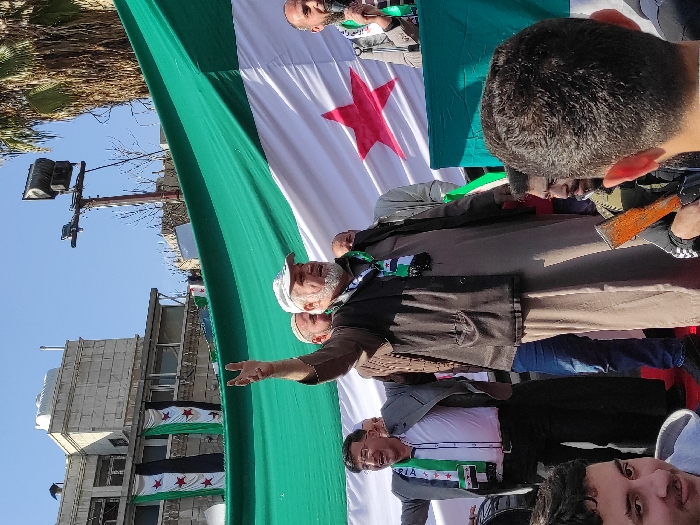 The image captures an outdoor gathering in Al-Qanawat Municipality, Syria, under a clear blue sky. A large banner, approximately 10 meters wide, displaying the Syrian opposition flag (green top stripe, white middle stripe with three red stars, black bottom stripe) forms the background. Two smaller, vertically hung Syrian opposition flags are visible on a building in the upper left background.
In the central foreground, a middle-aged male with a grey beard and a white cap, wearing a dark brown jacket over a light brown tunic, raises his right hand in a V-sign. He has a dark lanyard around his neck. To his immediate right (viewer's left), a man in a grey suit jacket and white shirt, adorned with a Syrian opposition flag scarf, smiles broadly with both arms slightly raised. A black drum is partially visible next to him. To the central figure's immediate left (viewer's right), a bearded man in a grey jacket holds a microphone to his mouth, also wearing a Syrian opposition flag scarf.
Approximately five additional male figures are partially visible in the mid-ground, either behind or to the sides of the central figures. One man in the lower right foreground is seen from behind, obscuring part of the scene. Directly behind him, another male holds a rifle with a distinct orange-brown foregrip and stock. The environment includes a multi-story building with multiple windows in the upper left background and a utility pole topped with a large floodlight in the upper right, with sparse tree foliage and a palm tree visible behind it.