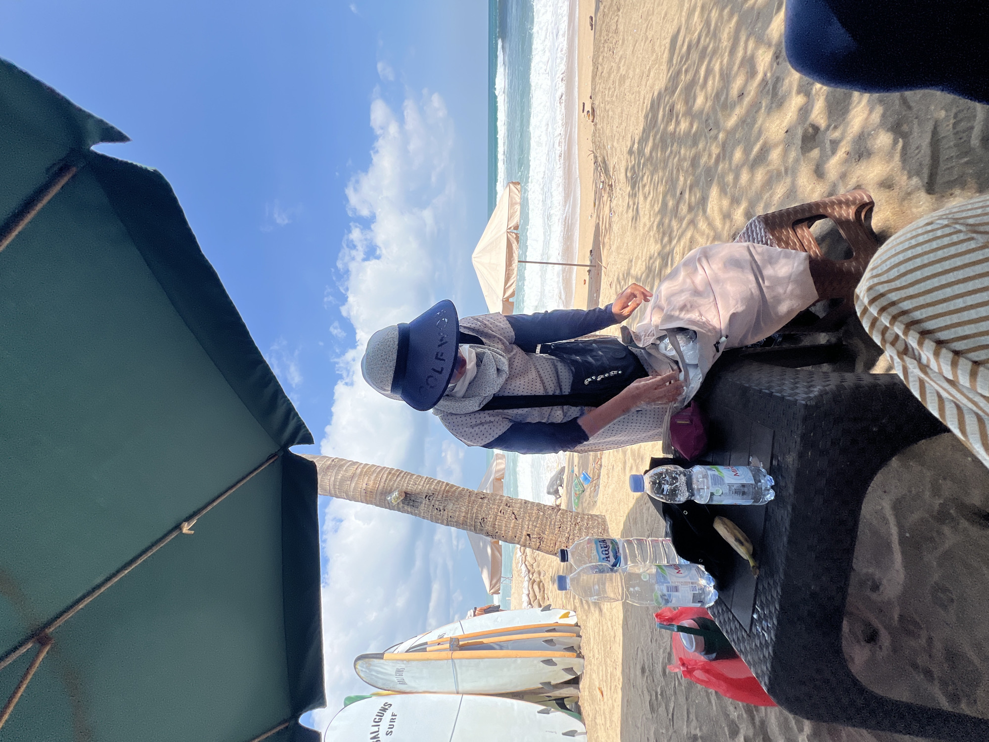 A person wearing a hat and hijab is standing behind a table on a sandy beach in Kuta, Indonesia. The person is reaching into a bag. On the table are several bottles of water, a banana, and a red bag. In the background, there are surfboards, palm trees, and the ocean with waves. A large green umbrella provides shade.