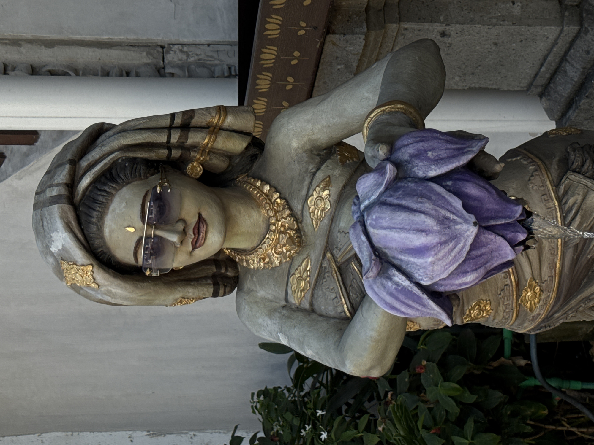 A close-up, eye-level medium shot captures a weathered, stone statue of a woman with green skin, wearing a large, draped headpiece and ornate gold jewelry, including a necklace, earrings, and armbands. She is also sporting modern aviator sunglasses, adding a contemporary touch to the traditional figure. The statue's hands cradle a large, purple lotus flower, which appears to be a water spout, as a thin stream of water is visible near the bottom of the flower. The background consists of a textured, pale wall on the left, lush green foliage, and a stone pillar and carved architectural elements on the right, suggesting an outdoor setting in a temple or garden. The lighting indicates it is daytime, with soft shadows suggesting an overcast sky or diffused sunlight. The statue's serene expression and the water feature imply it is a decorative element, possibly a fountain, in a location like Ubud District, Indonesia, a region known for its rich cultural heritage and artistic traditions.