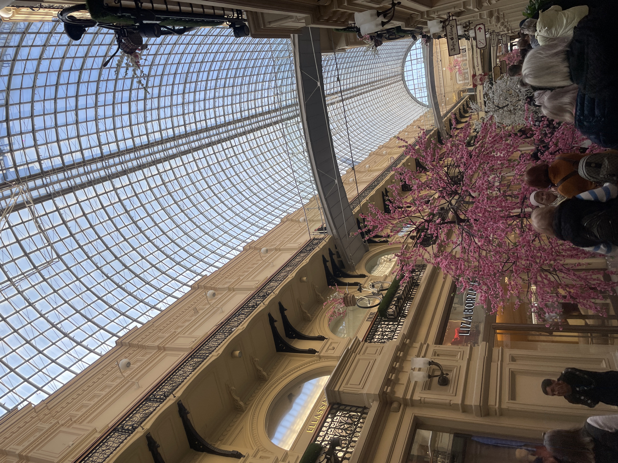The image depicts the interior of the GUM department store in Moscow, Russia. The store is characterized by a distinctive glass ceiling supported by a steel framework, which allows natural light to flood the building. The architecture features multiple levels with decorative moldings, arched windows, and wrought-iron railings. Mannequins are displayed on the upper floors. Artificial cherry blossom trees with pink flowers are a prominent decorative feature. People are present in the scene, seemingly shopping or passing through the store. The name "LIZA BORZILOVA" is visible on a storefront. The photograph has been taken from a low angle, looking upwards towards the ceiling and upper levels of the store.