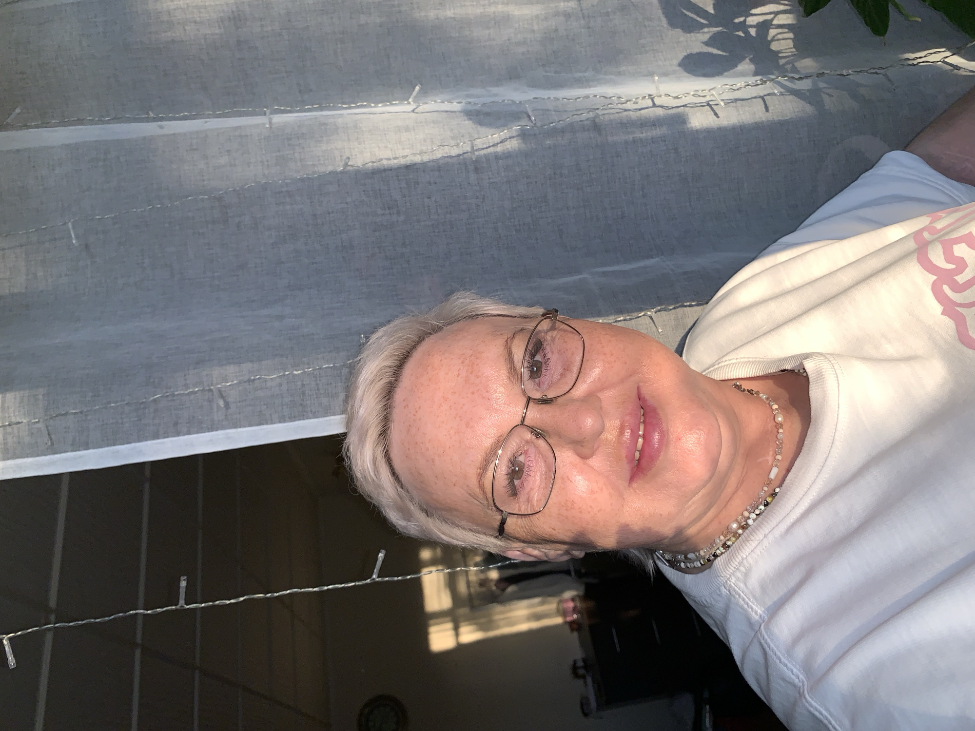 A woman with short white hair and glasses is wearing a white shirt and a beaded necklace. She is smiling. Behind her is a sheer white curtain, and underneath that, a room with a dark table and a window. A string of lights hangs above the table. The location is unknown, but the prompt mentions the city of Chaikovsky, Russia.