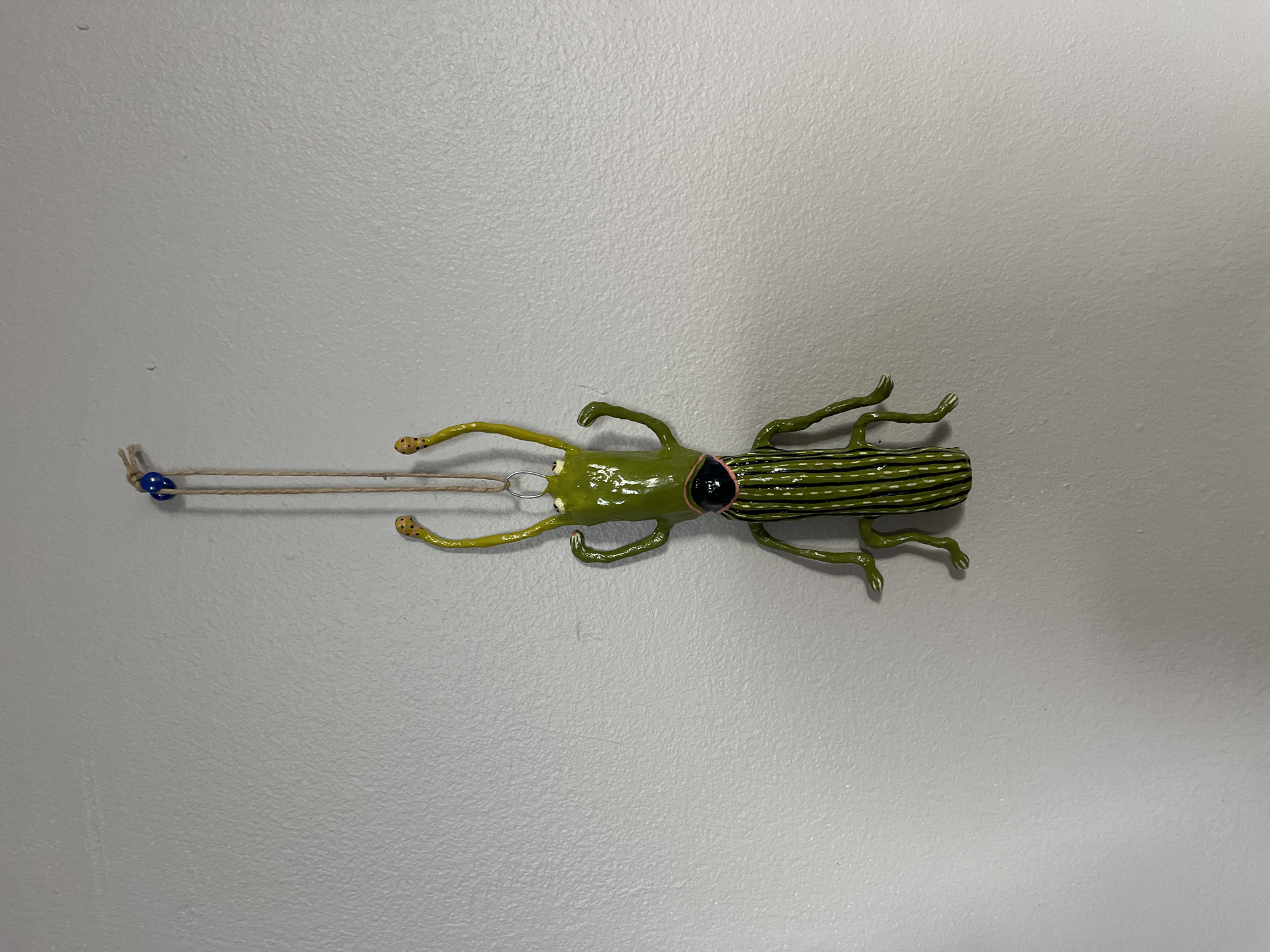 A painted metal sculpture of a green beetle is displayed horizontally on a textured, light gray wall. The beetle is oriented with its head to the left and its segmented abdomen to the right. The head is small and green, with black eyes. Its antennae are thin and yellow, and it has two thin, yellow arms that extend forward. The beetle's body is a bright green, and its abdomen is decorated with thin, dark green or black stripes. Four thin, green legs are visible, two on each side, extending downward from the body.
A thin, tan string is attached to the beetle's head. The string extends to the left, and a small, blue bead is threaded onto it. The string is taut, suggesting the beetle is hanging or suspended. The background is a plain, light gray wall with a slightly textured surface, typical of interior walls. The lighting appears to be artificial, casting a soft, even illumination on the scene. There is no indication of the time of day, weather, or specific location beyond the general context of being indoors. No text is visible in the image.
