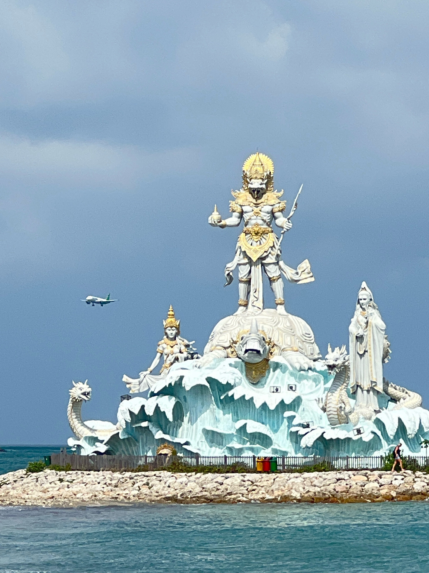 The image shows the monumental Garuda Wisnu Kencana (GWK) statue in Kuta, Bali, Indonesia. The statue depicts Lord Vishnu riding the mythical Garuda bird. In the foreground, the turquoise waters of the ocean are visible, with a rocky embankment in front of the statue. The statue itself is large and intricately detailed, featuring golden accents on a white structure. Several smaller statues surround the main monument, including what appear to be mythological creatures. An airplane is seen flying in the distance in the sky. The sky is cloudy. A person is walking along the rocky embankment.