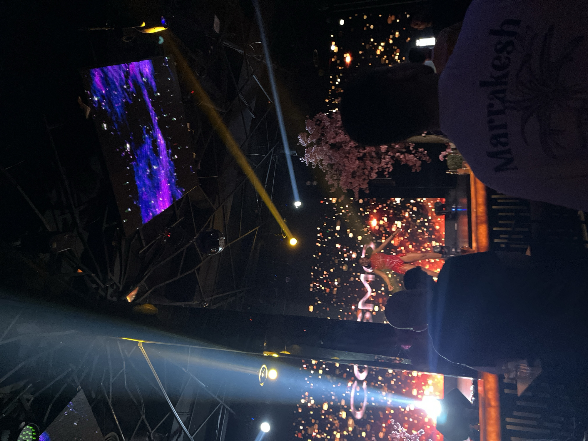 The image shows a crowded indoor venue in Plovdiv, Bulgaria, likely a nightclub or event space. Stage lighting illuminates large video screens displaying abstract graphics and figures, with one screen showing a performer in a red dress. A person in the foreground wears a white shirt with the word "Marrakesh" printed on the back. The scene is lively, with attendees viewing the stage performance. Structural elements with a mirrored, geometric design are visible.