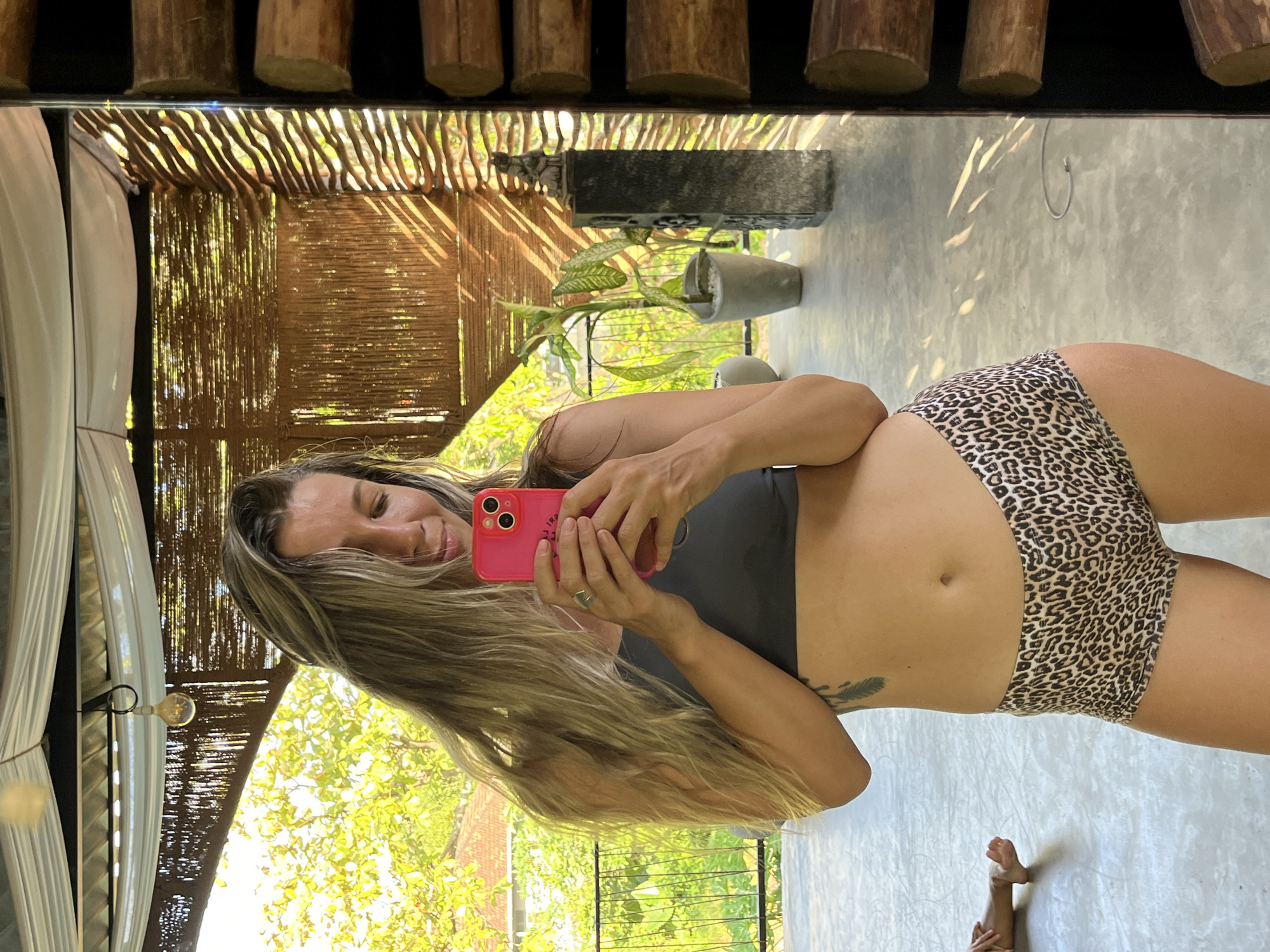 A woman stands in front of a mirror, taking a selfie. She is wearing a gray sports bra and leopard-print shorts. Her hair is long and blonde, and she has a tattoo on her left side. The background appears to be an outdoor patio with bamboo walls and a thatched roof. Sunlight filters through the bamboo, creating patterns on the floor. There are plants in pots and a statue on a pedestal. In the reflection of the mirror, another person's feet are visible on the floor. The scene is likely in Weligama, Sri Lanka, given the tropical vegetation and architectural style.