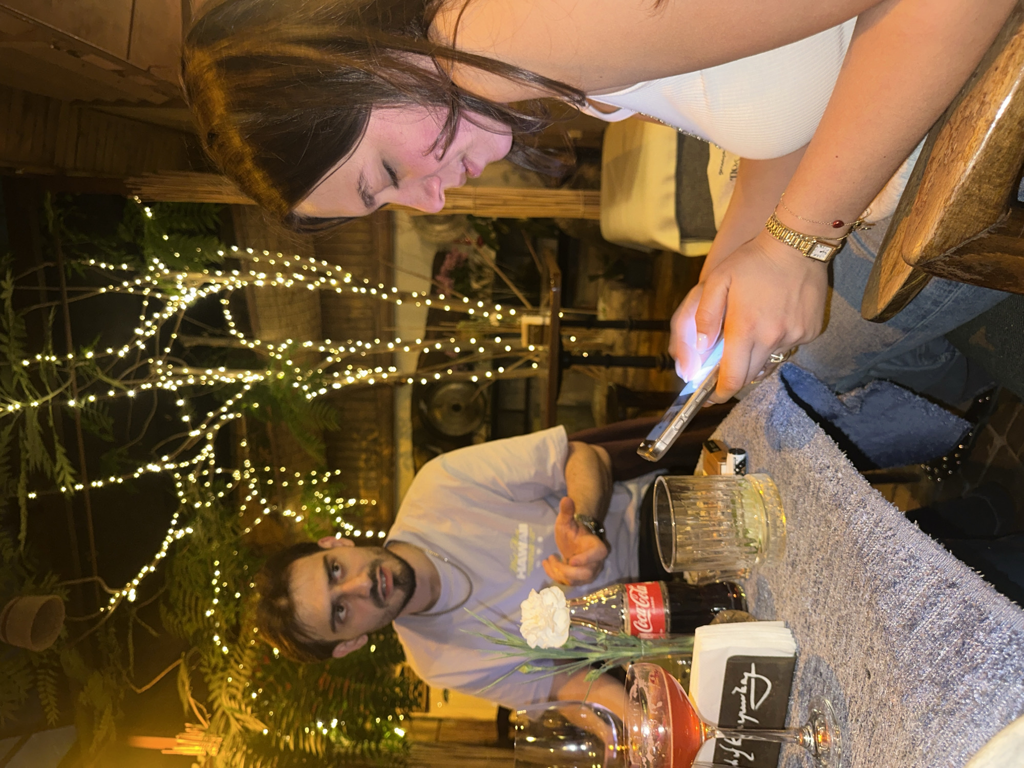 The image captures an indoor setting, likely a restaurant, in Yerevan, Armenia. A woman with long brown hair is looking down at her cell phone. She is wearing a white tank top and blue jeans. A gold watch is visible on her wrist. A man with short brown hair and a beard is seated at the table, facing the camera. He is wearing a light purple shirt and a silver necklace.
On the table is a light blue fabric tablecloth. Several items are arranged on the table, including a clear drinking glass, a bottle of Coca-Cola, a white carnation, a wine glass filled with a red liquid, and a stack of black and white napkins.
Strings of miniature white lights provide illumination and decorate the background. Wooden structures and greenery are also visible in the background, suggesting a rustic or outdoor-themed interior design.